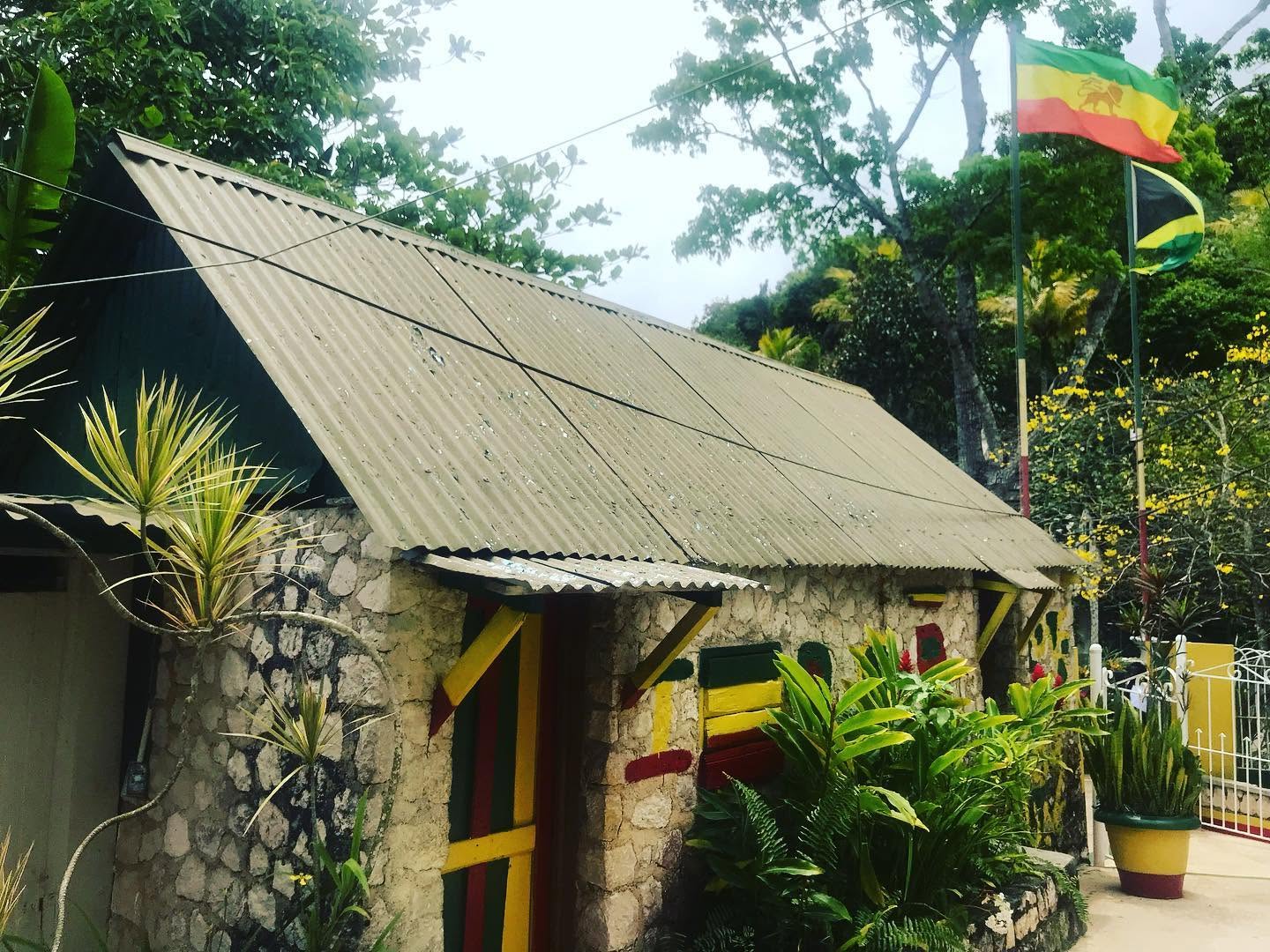 Stone house with a sloped roof, surrounded by greenery and flags, including a yellow, red, and green flag, in a garden setting.