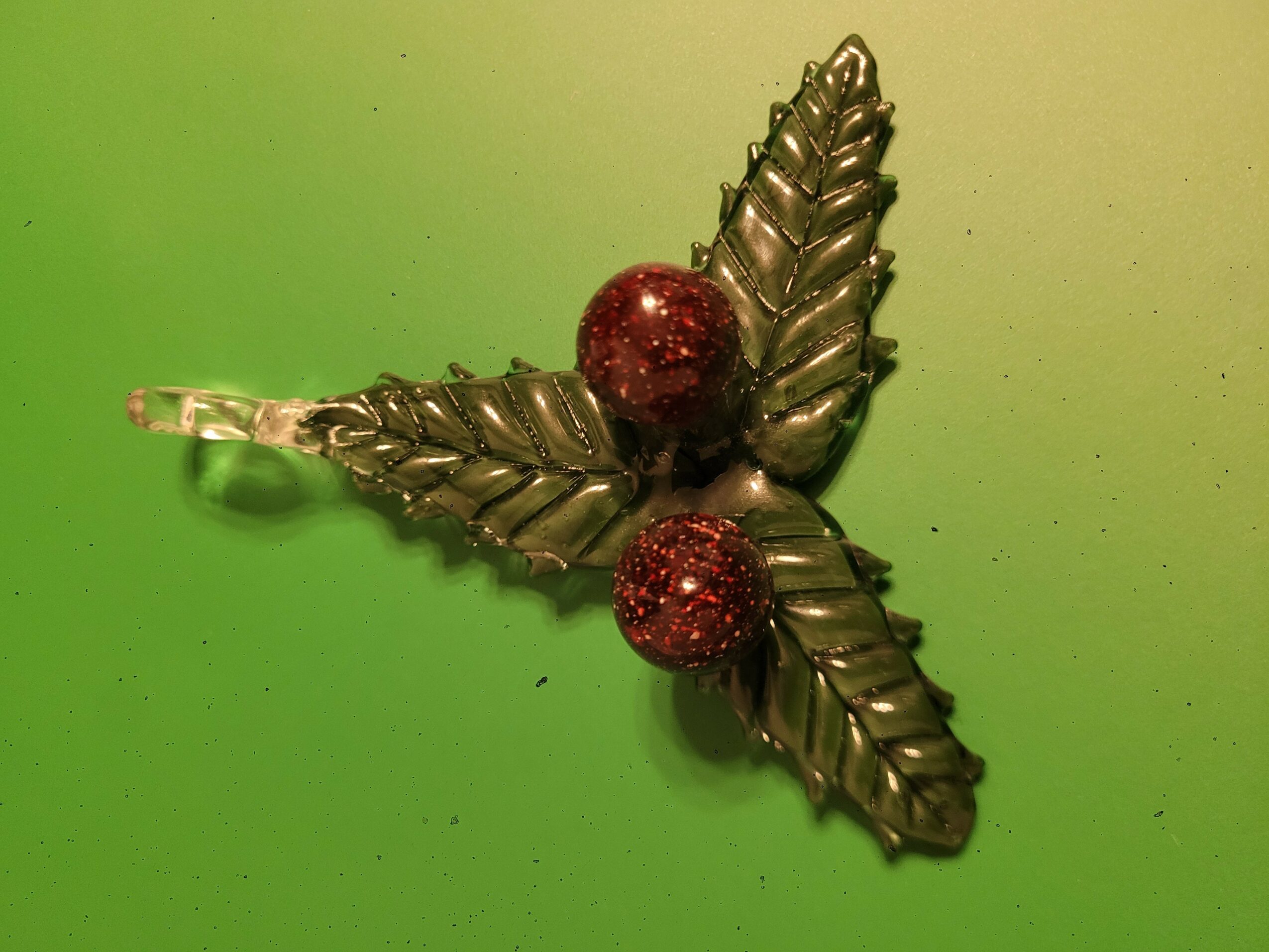 Decorative ornament with three metallic leaves and two red glittery berries on a green background.
