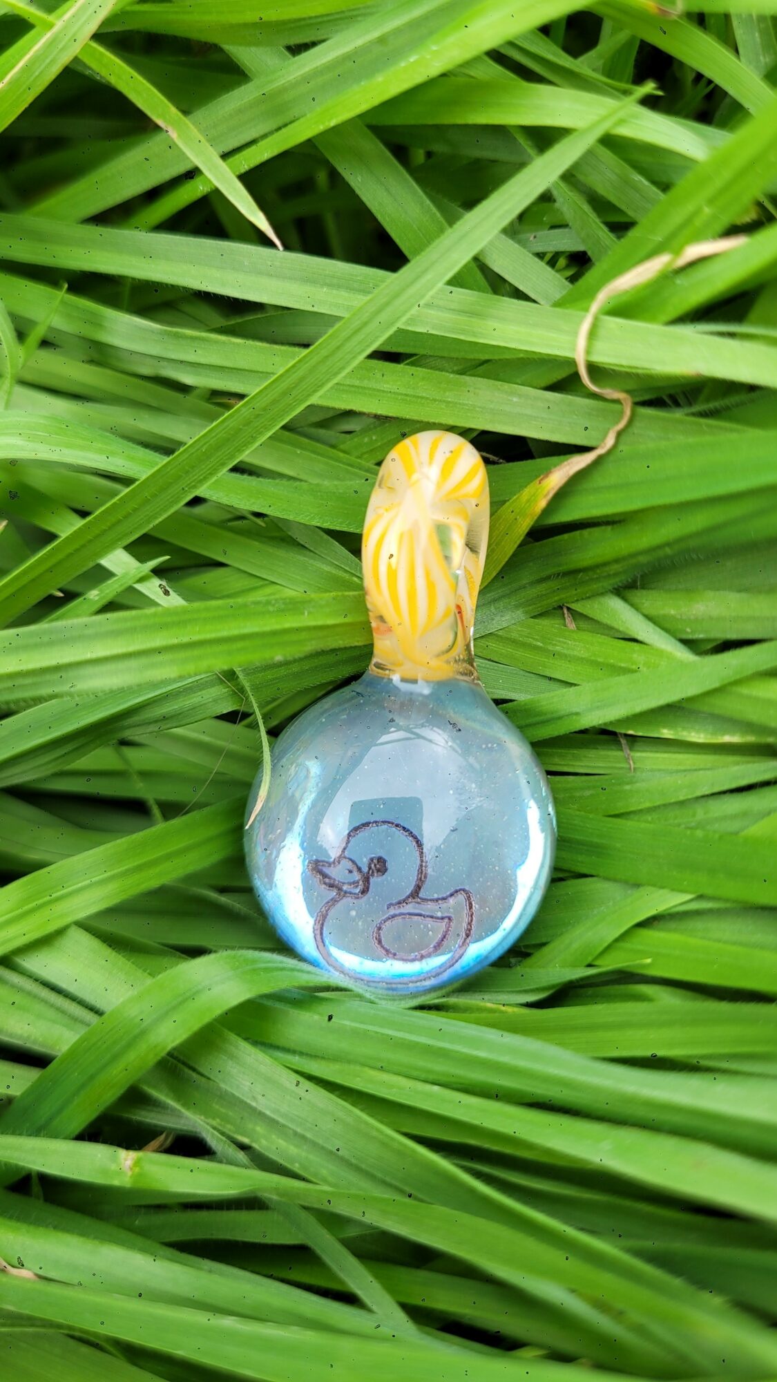 Glass pendant with a yellow and white top, containing a small blue whale illustration, resting on green grass