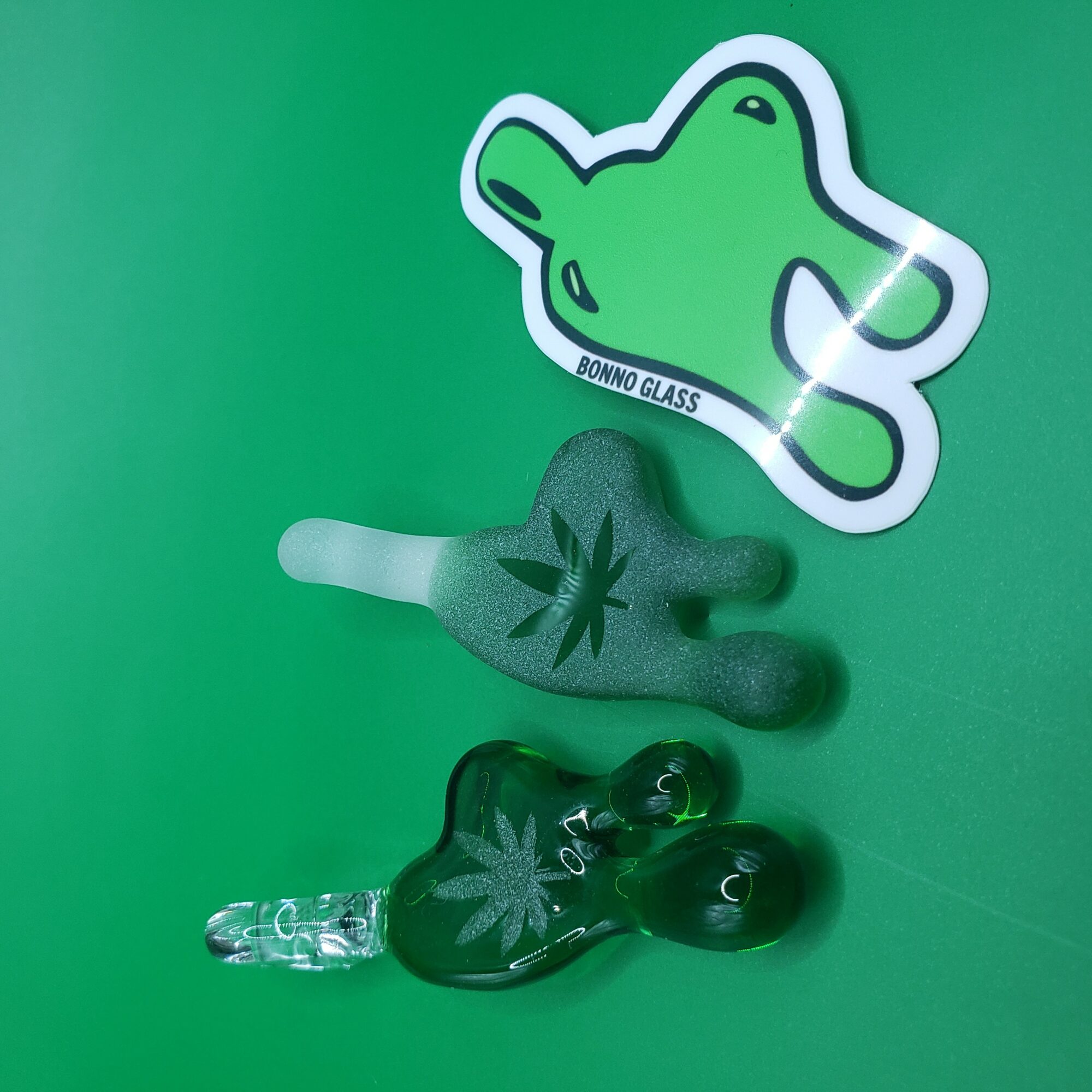 Three glass objects with marijuana leaf designs and a green bear-shaped sticker on a green background.