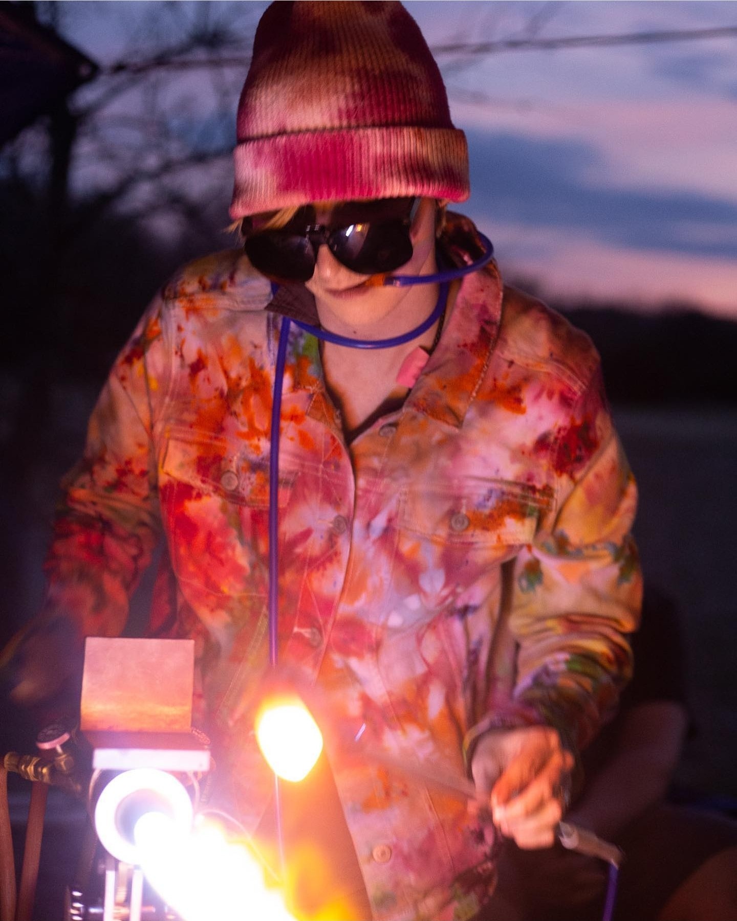 Child wearing a colorful jacket, beanie, and sunglasses, holding a lit object outdoors at dusk.