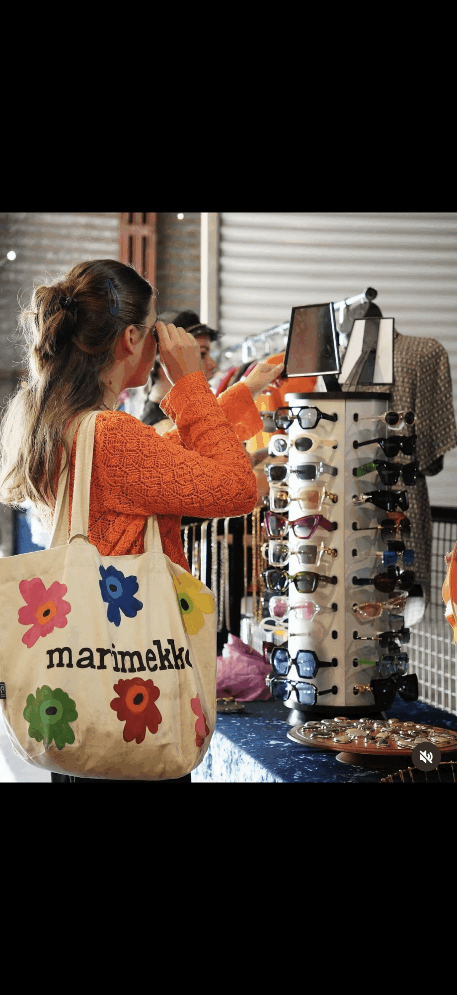 Person with long hair in an orange sweater looking at a tablet at a market stall with sunglasses and accessories.
