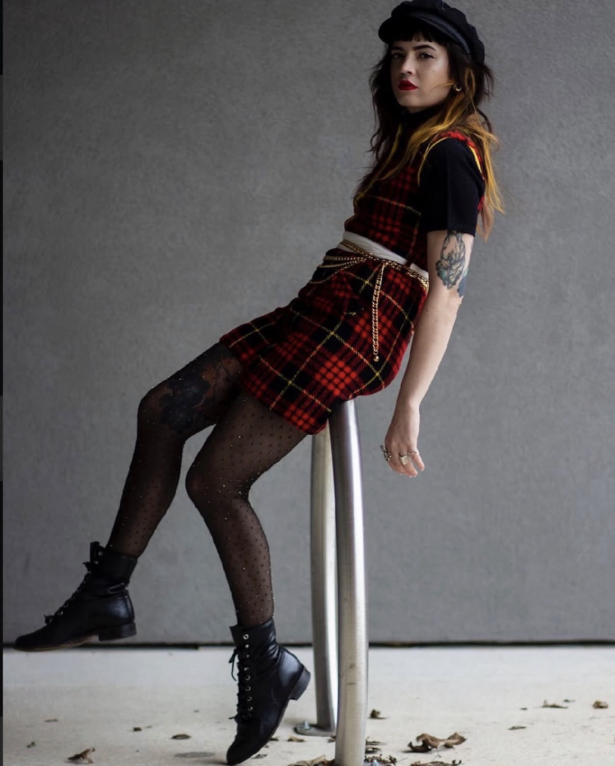 Young woman with dark hair and tattoos sitting on a stool against a plain gray background, wearing a plaid skirt, black top, and boots.