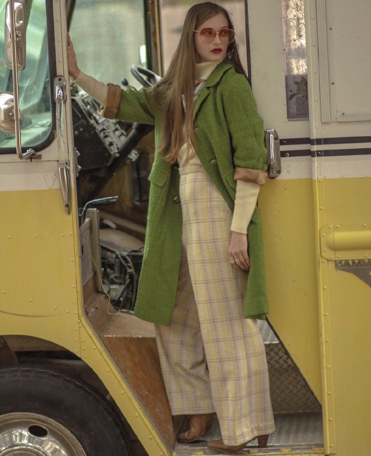 Woman with long hair and glasses standing in a yellow vehicle doorway, wearing a green coat and plaid pants.