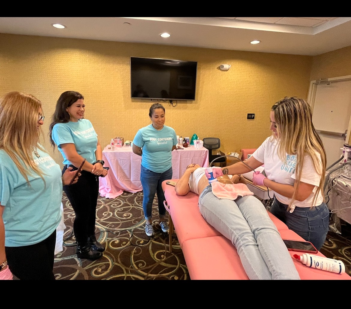 Four women and a girl in a room with a woman lying on a medical table, receiving a procedure from another woman. A TV is on the wall.
