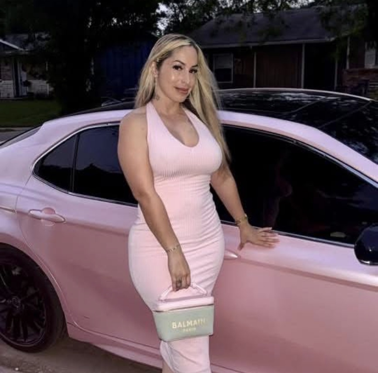 Woman with long blonde hair in pink dress holding a purse, standing next to a pink car outdoors.