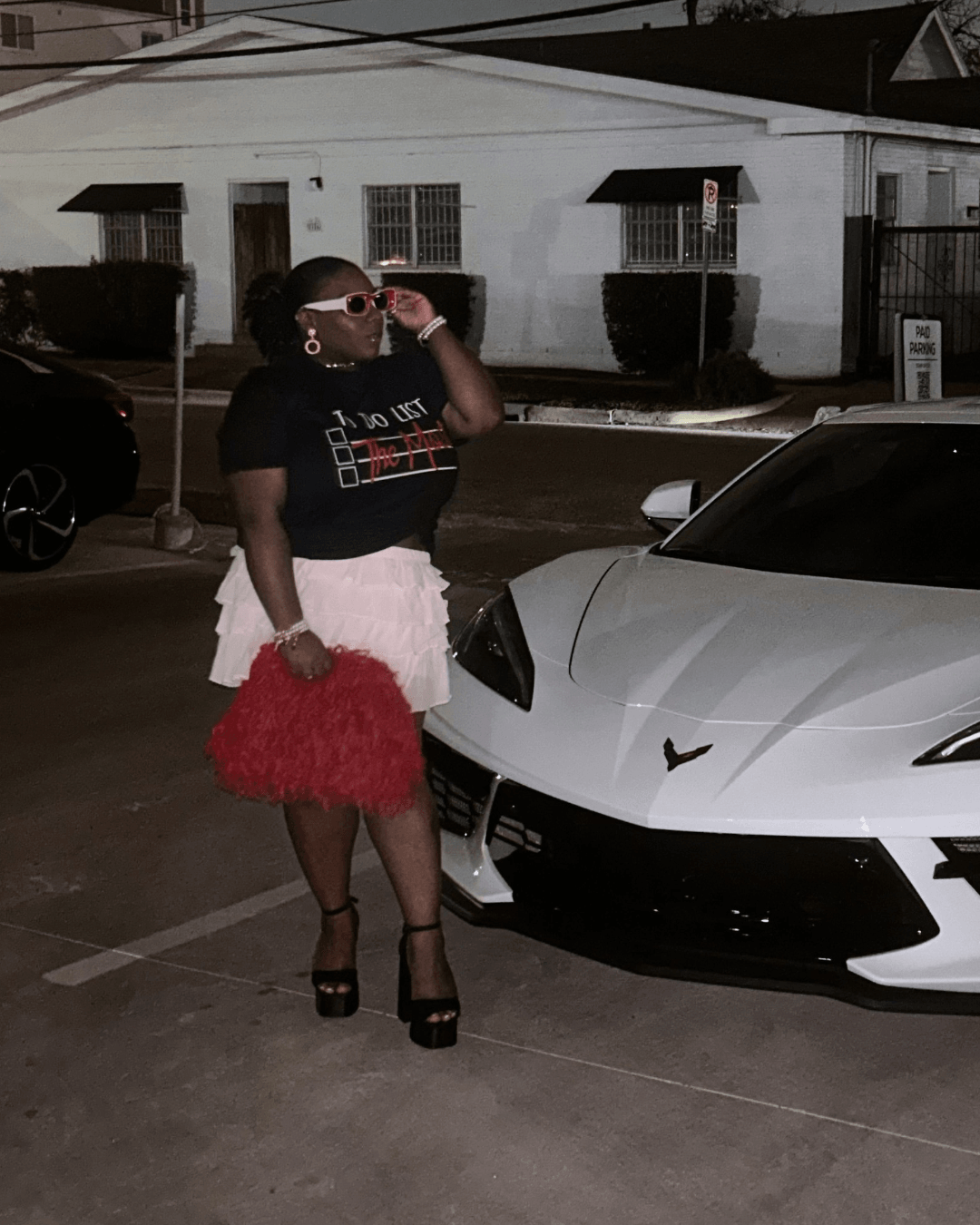 Person standing next to a white sports car, holding a red fluffy bag, wearing black heels and sunglasses, in a parking lot.