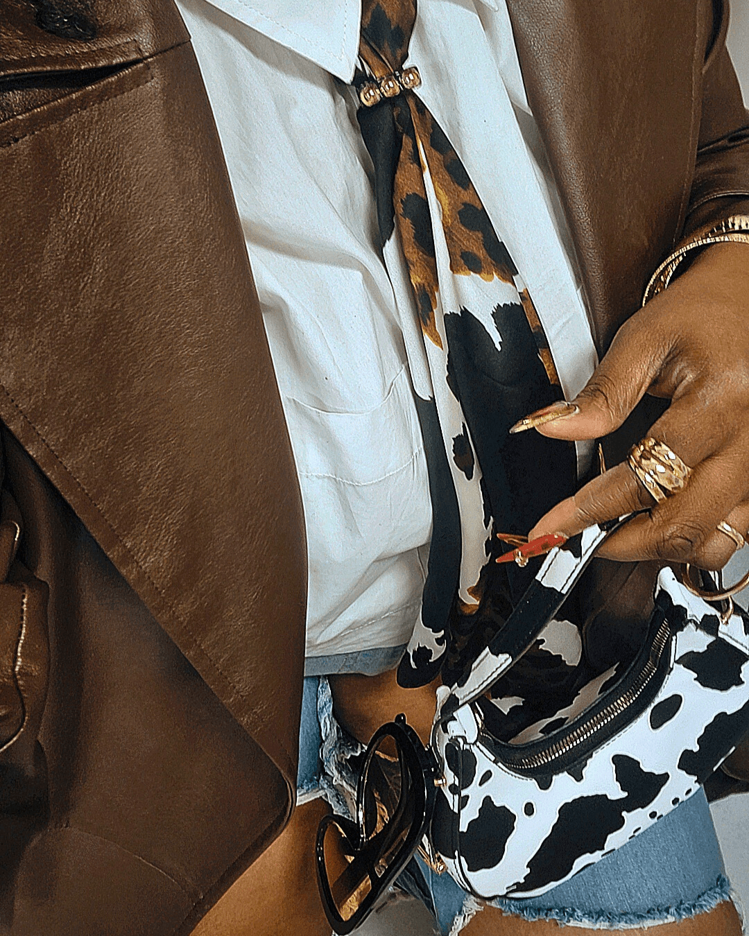 Person wearing a white shirt, leopard print tie, brown jacket, and holding a cow-print bag, with rings on fingers.