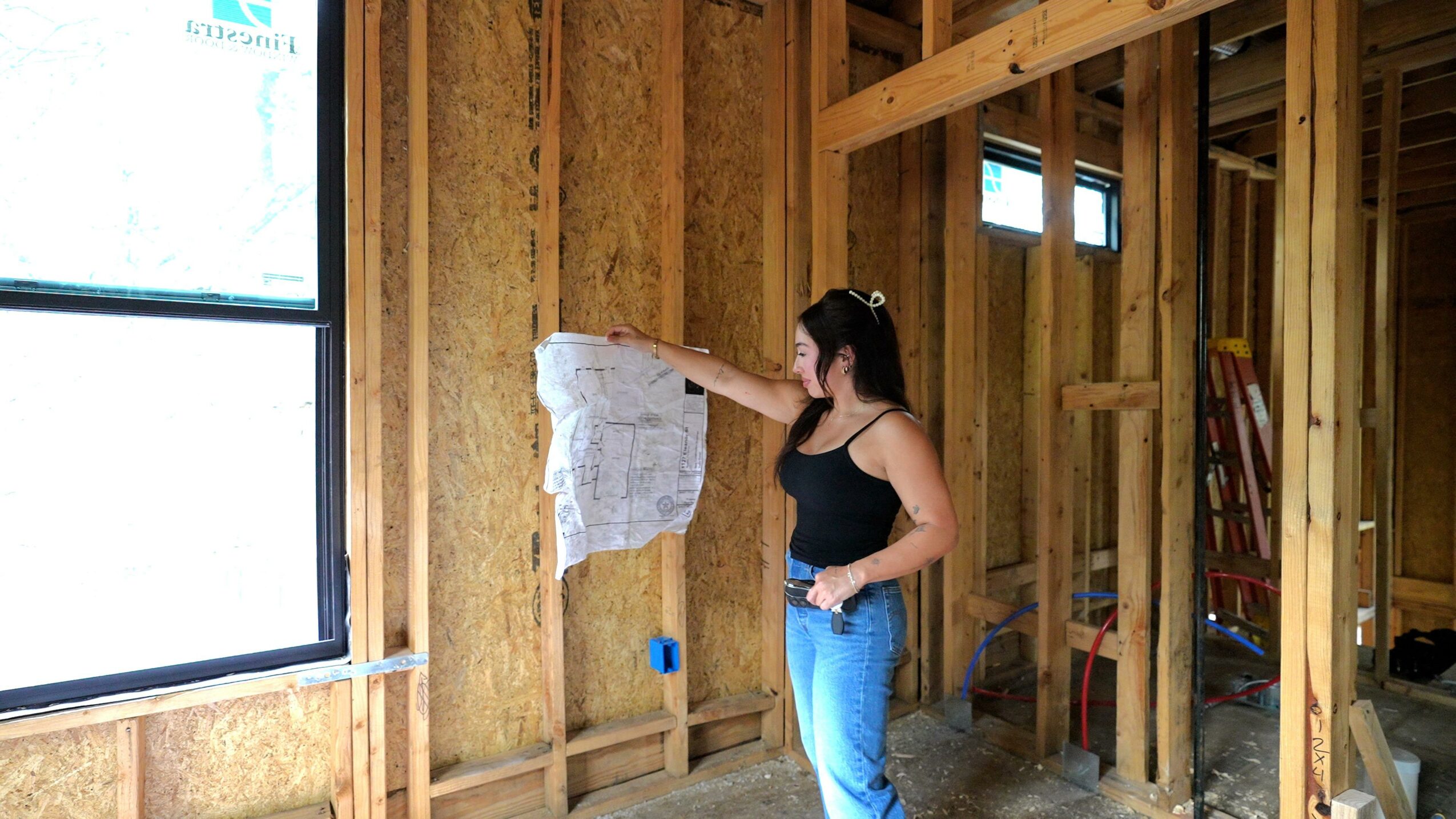 Woman inspecting a window in a wooden frame construction site, holding a cloth or paper. Interior with exposed wood studs.