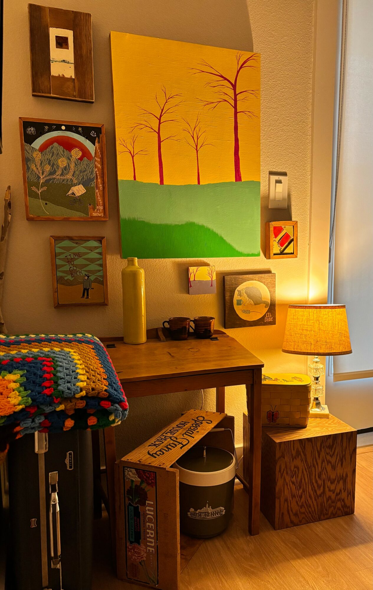 Interior corner with colorful paintings, a wooden table, a lamp, and a crocheted blanket, with a window on the right.