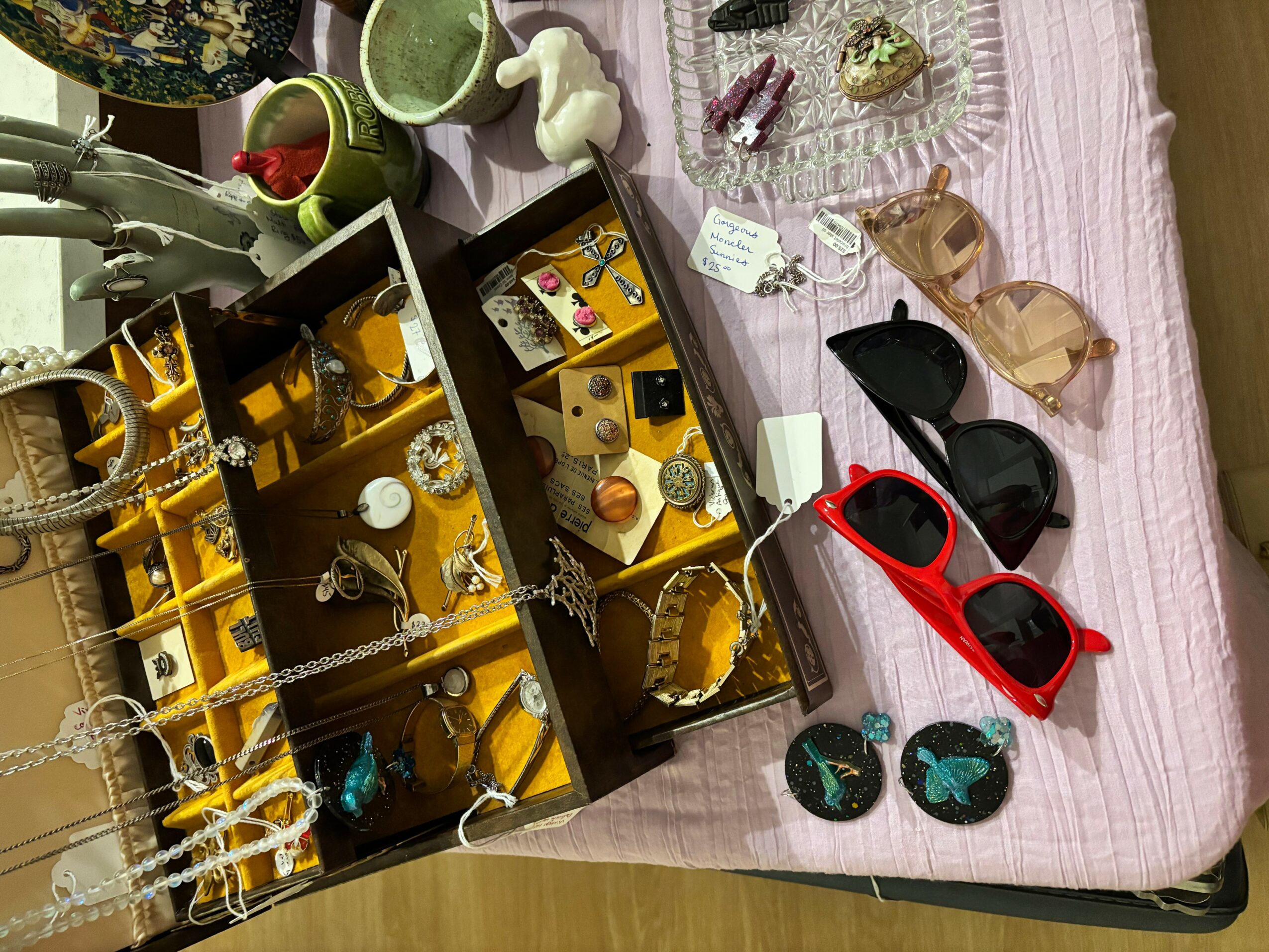 Jewelry display with necklaces, rings, and earrings, along with sunglasses and small accessories on a pink tablecloth.