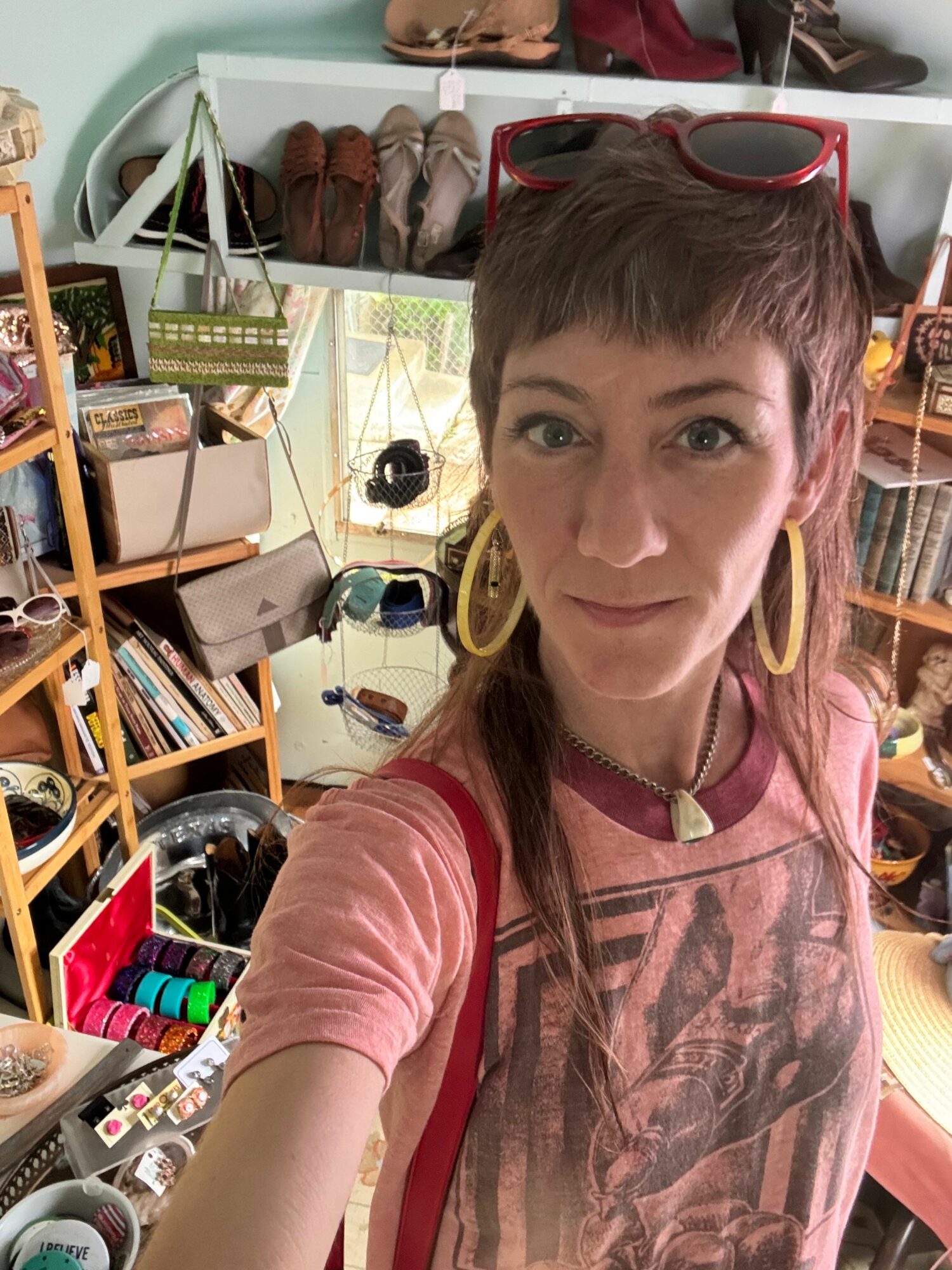 Woman with short hair and sunglasses on her head takes a selfie in a store, surrounded by shelves and products.