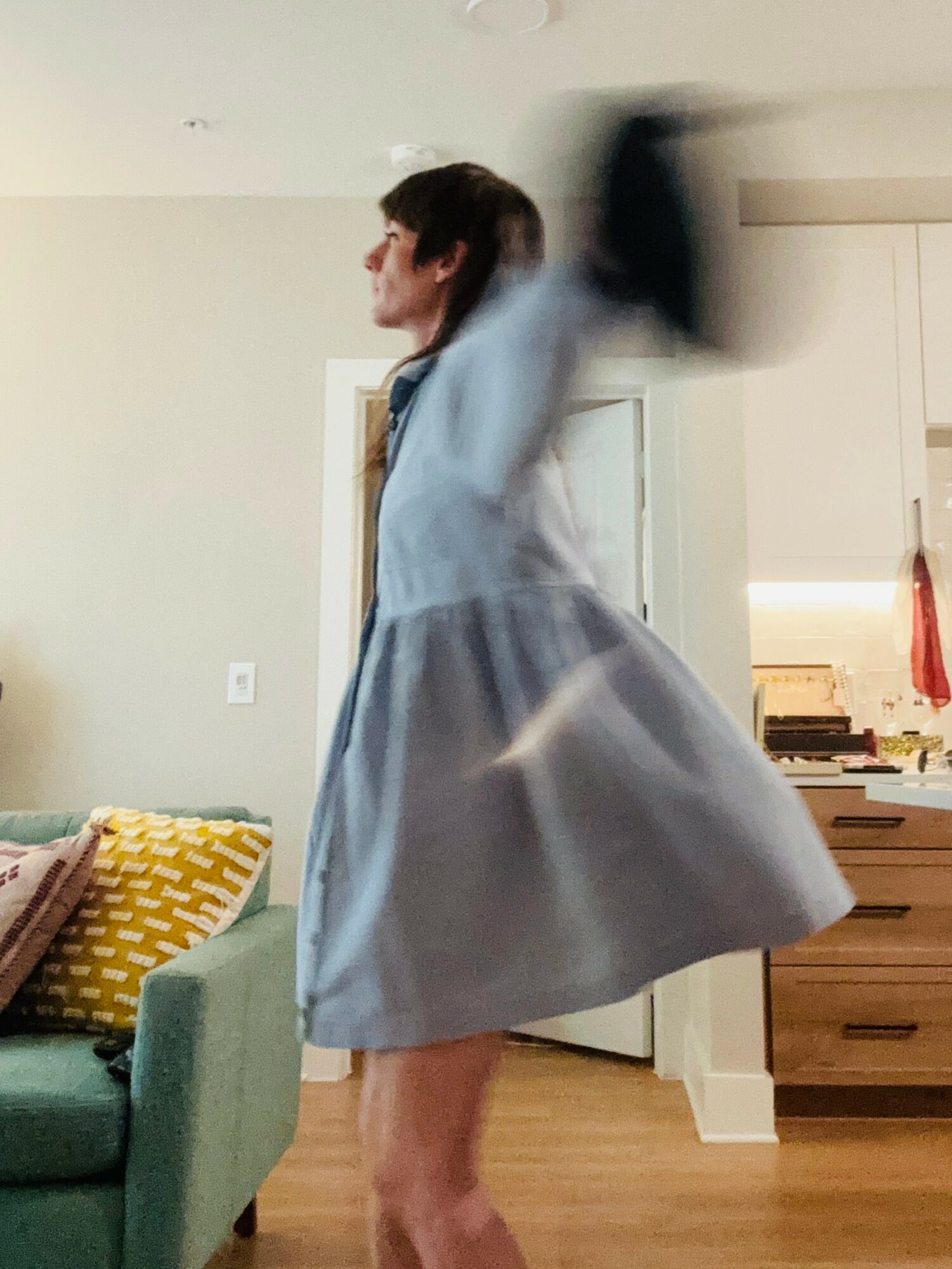 Person in a light blue dress spinning with arm raised, motion blur visible, in a room with a green couch and wooden furniture.