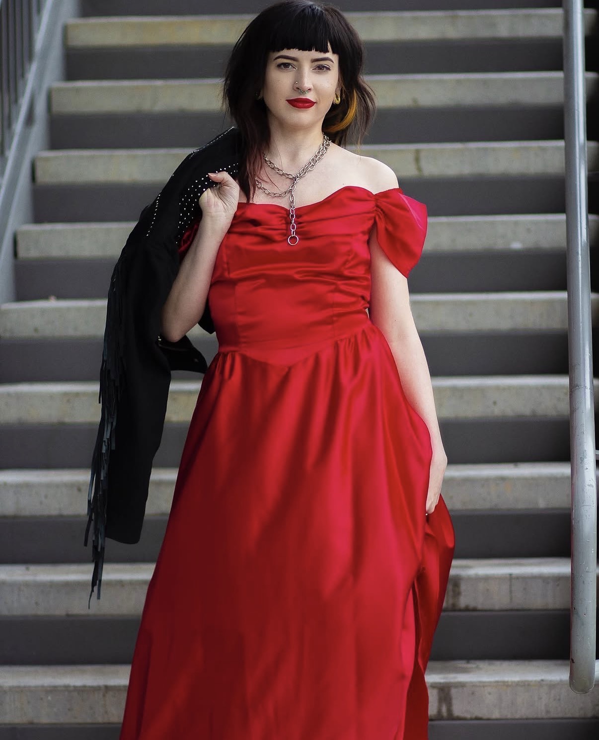 Woman in red dress standing on stairs, holding a black jacket over her shoulder, with dark hair and red lipstick.