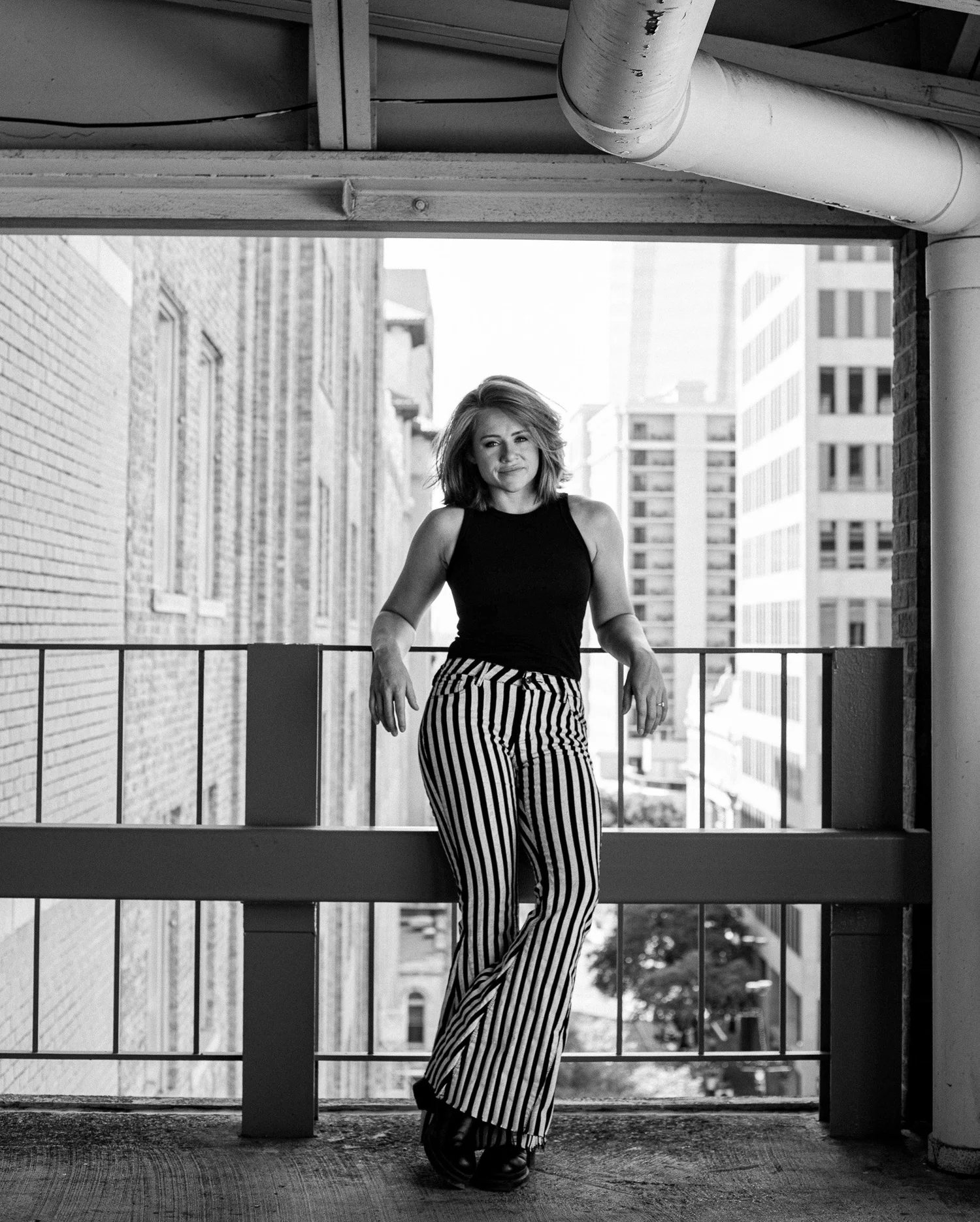 Woman standing on balcony with city buildings in background, wearing striped pants and a sleeveless top.
