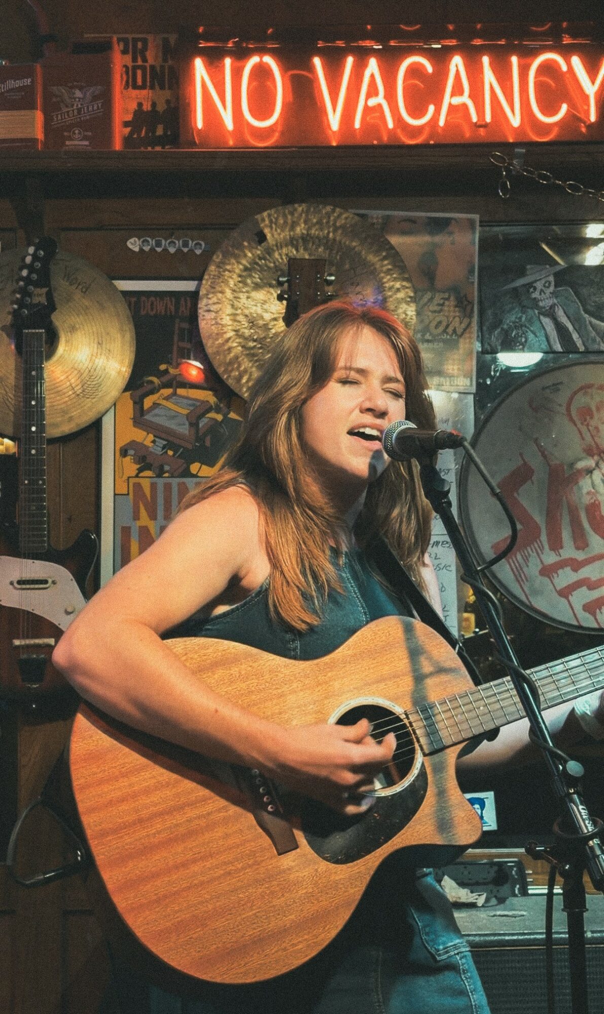 Woman singing and playing guitar in a bar with a neon 'No Vacancy' sign above.