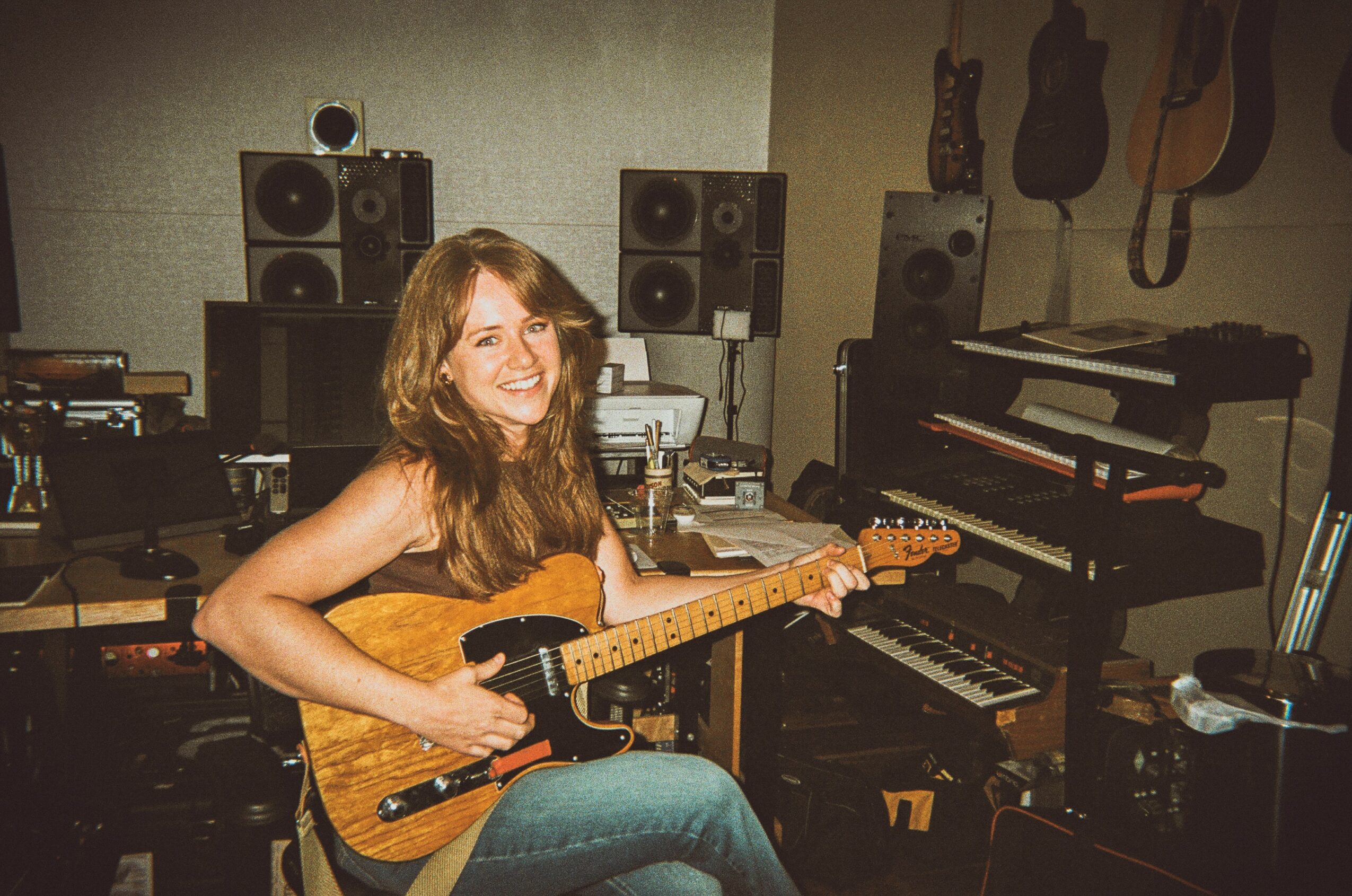 Woman with long hair playing an electric guitar in a music studio with speakers and guitars on the wall.