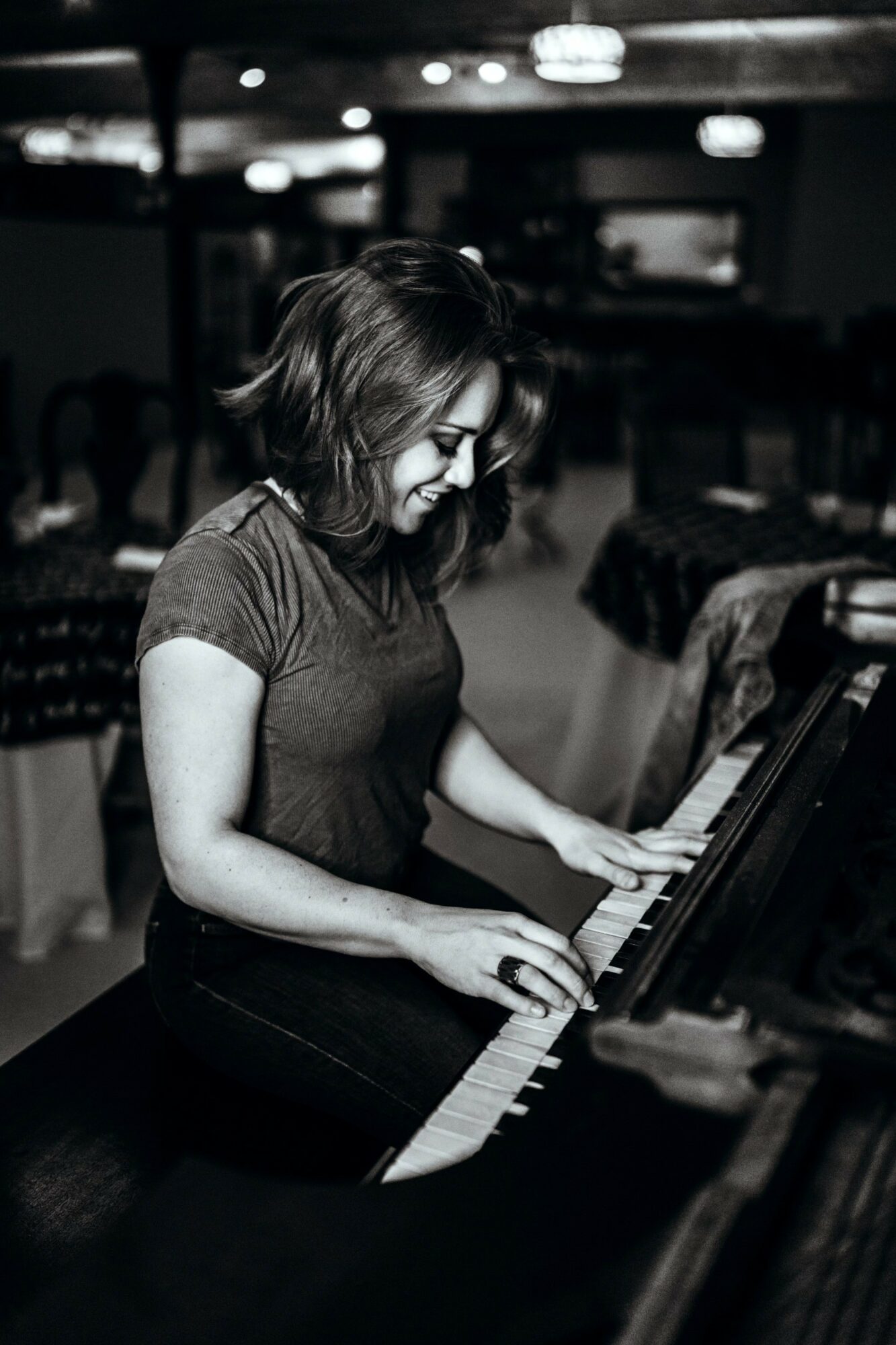 Woman playing piano, smiling, in a dimly lit room, with chairs and tables in the background.