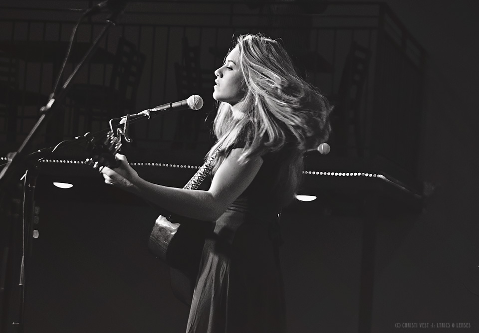 Woman singing into a microphone while playing a piano on stage, with her hair flowing, in black and white.