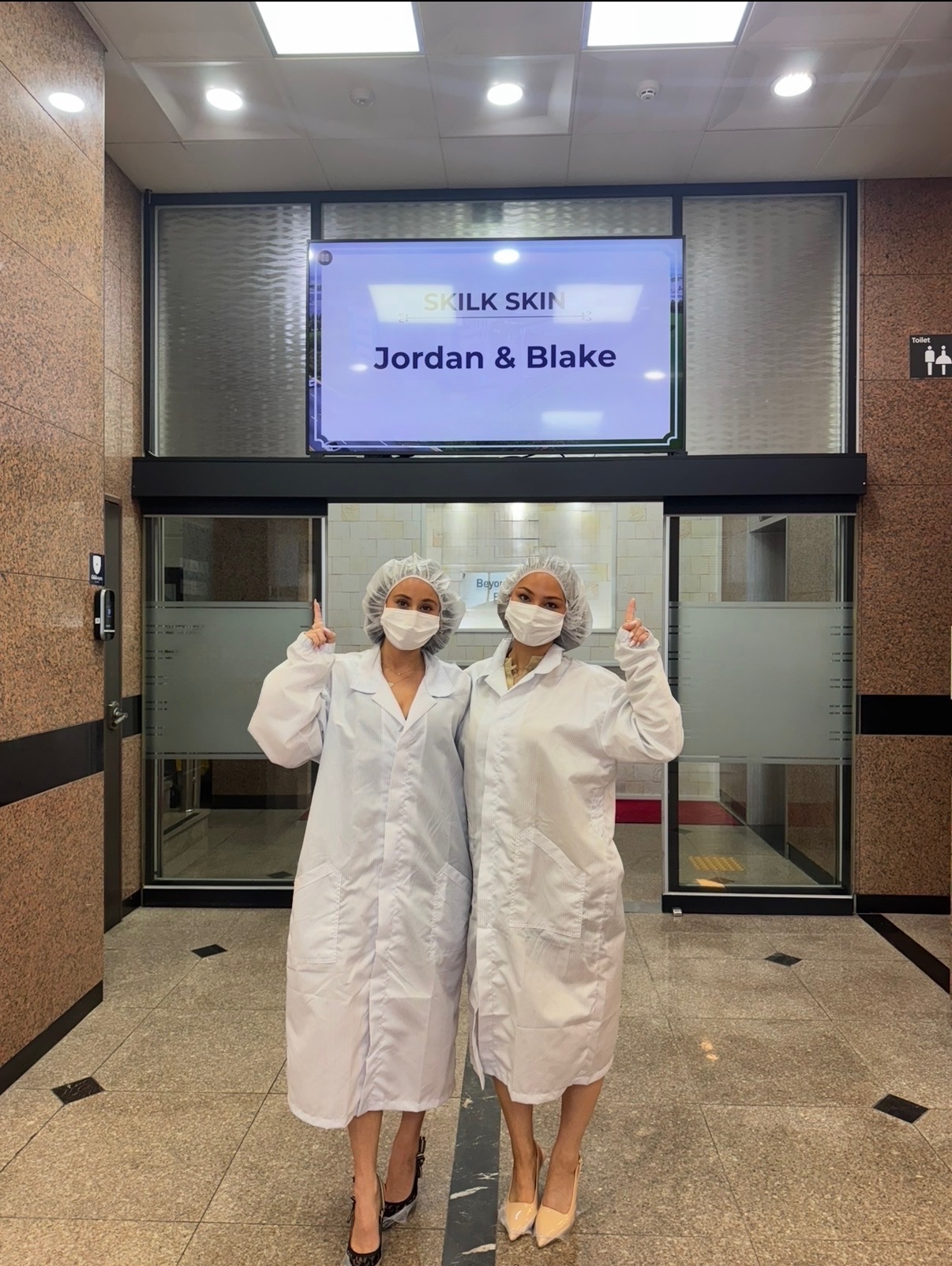 Two healthcare workers in white coats and masks stand in front of an elevator, giving thumbs up.