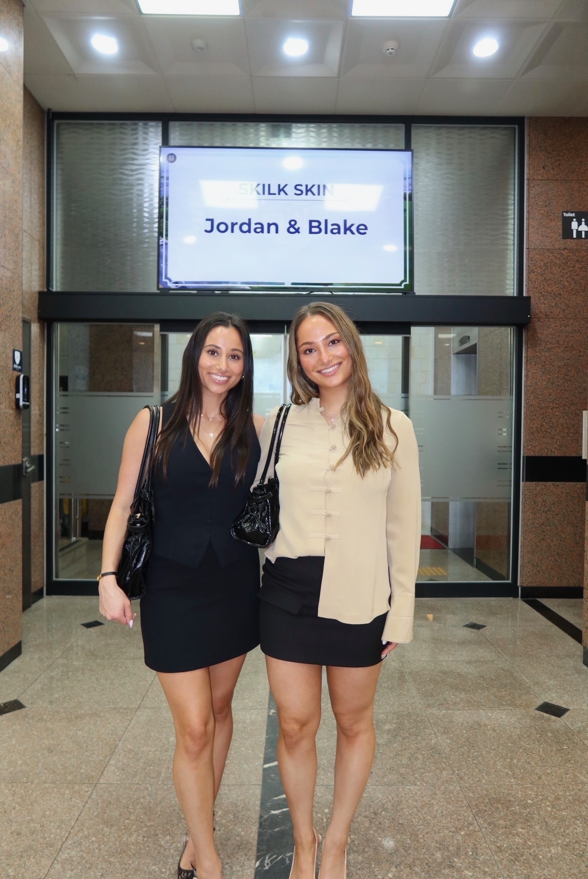 Two young women stand in front of an elevator with a digital sign above displaying names, smiling at the camera.