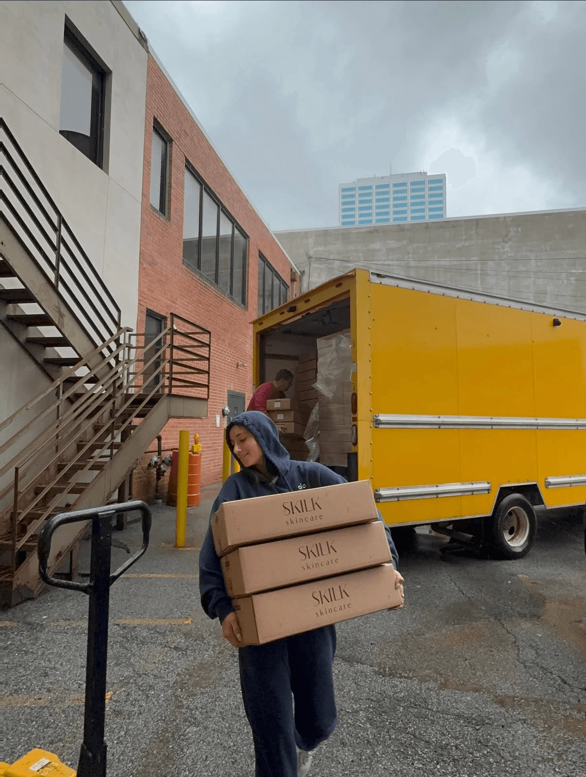 Person carrying three boxes labeled 'SUNK' standing near a yellow truck in an urban area with buildings and cloudy sky.