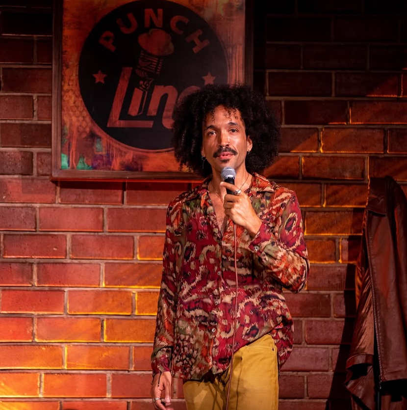 Person with curly hair holding a microphone on stage, brick wall background, comedy club sign behind them.