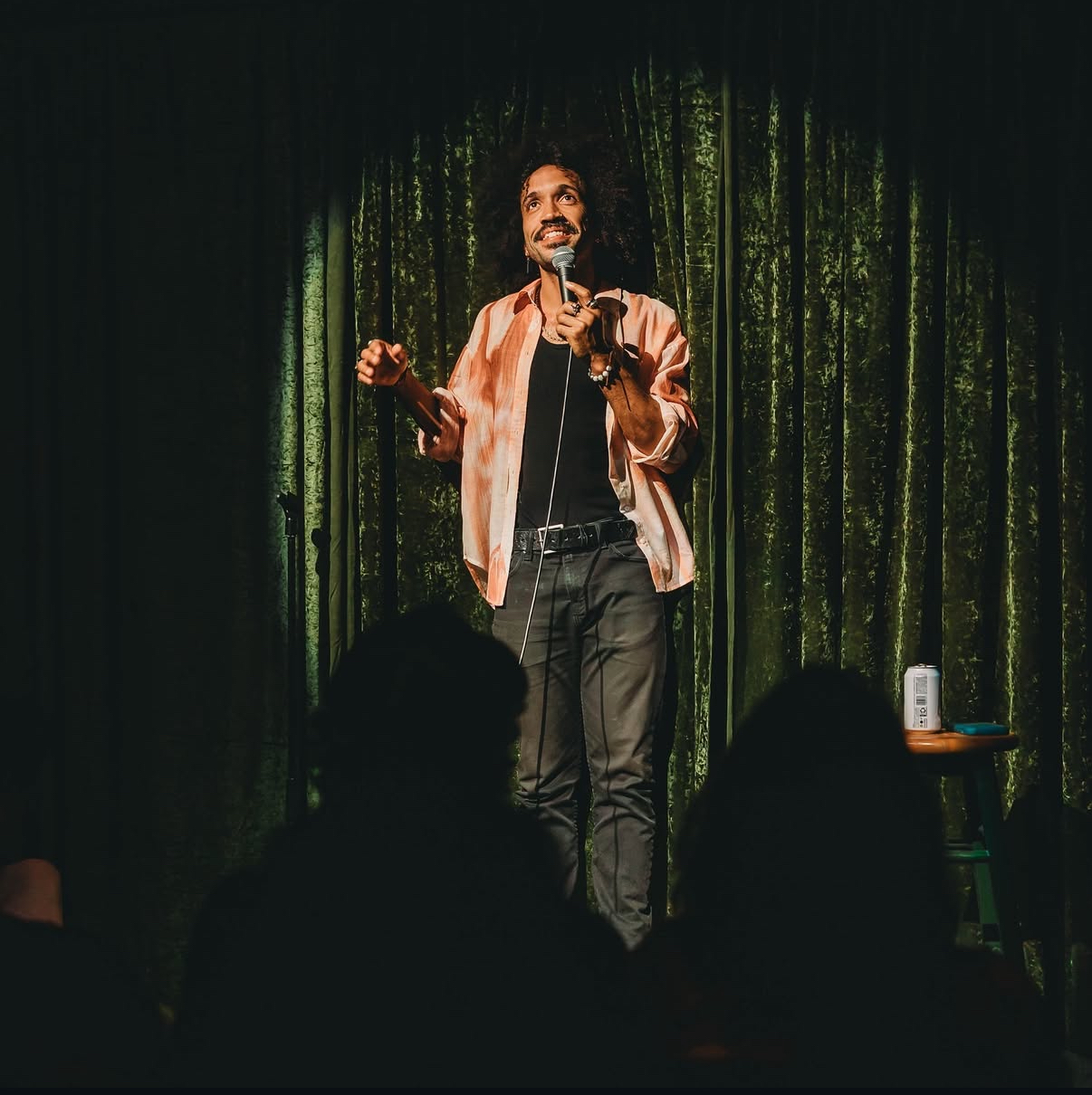 Person performing stand-up comedy on stage with green curtain background, holding microphone, audience silhouettes visible, small table with a drink.