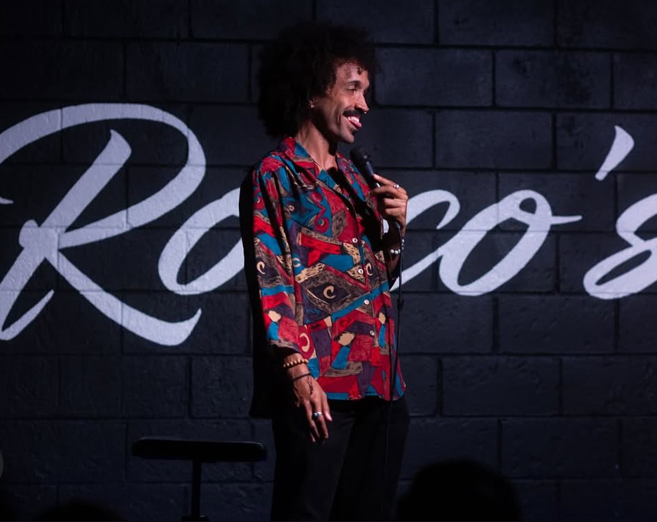 Person with curly hair wearing a colorful patterned shirt, standing on stage with a dark background and white text, smiling.