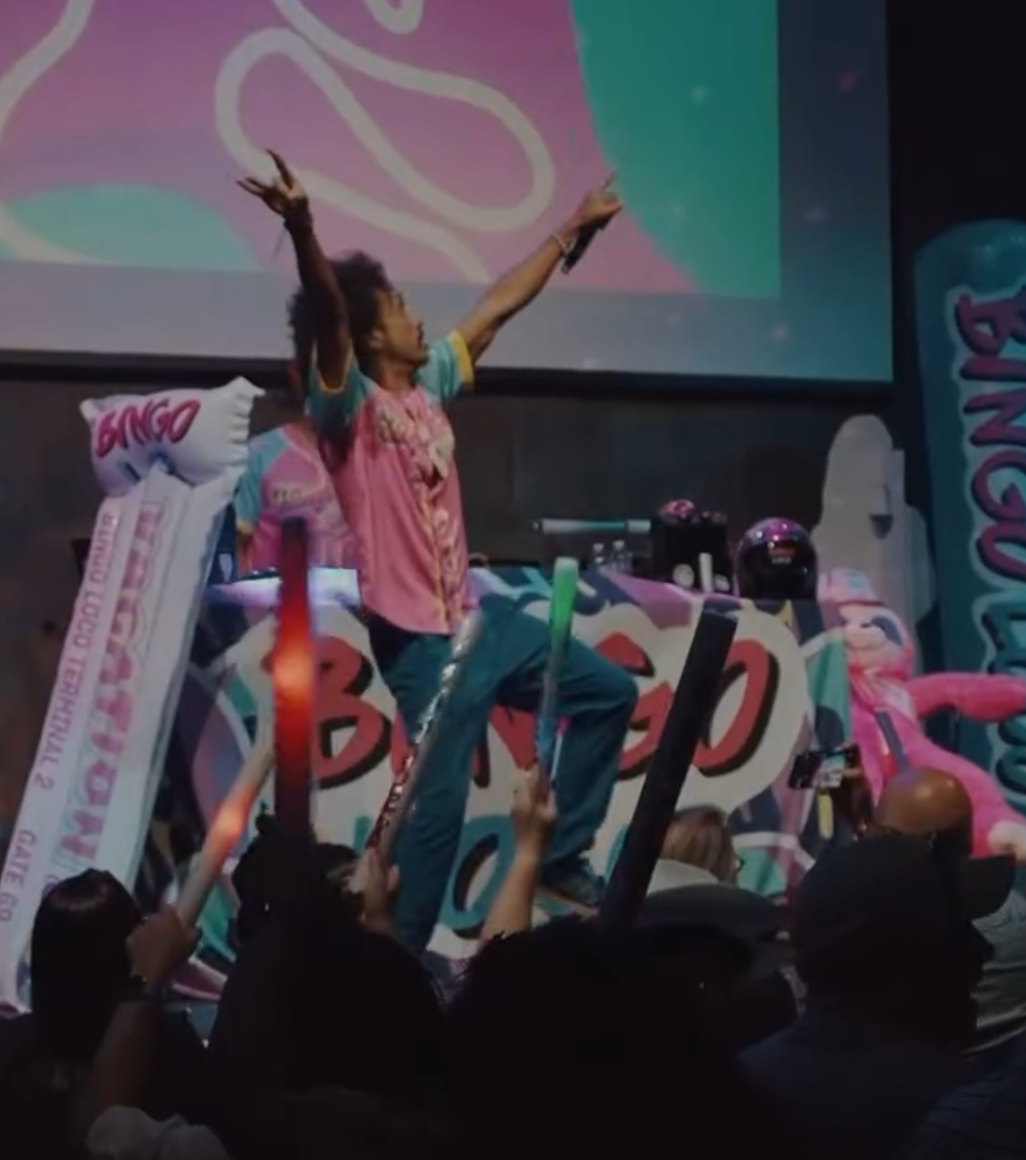 Person on stage with arms raised, colorful background, audience watching, pink and blue decorations, lively scene.