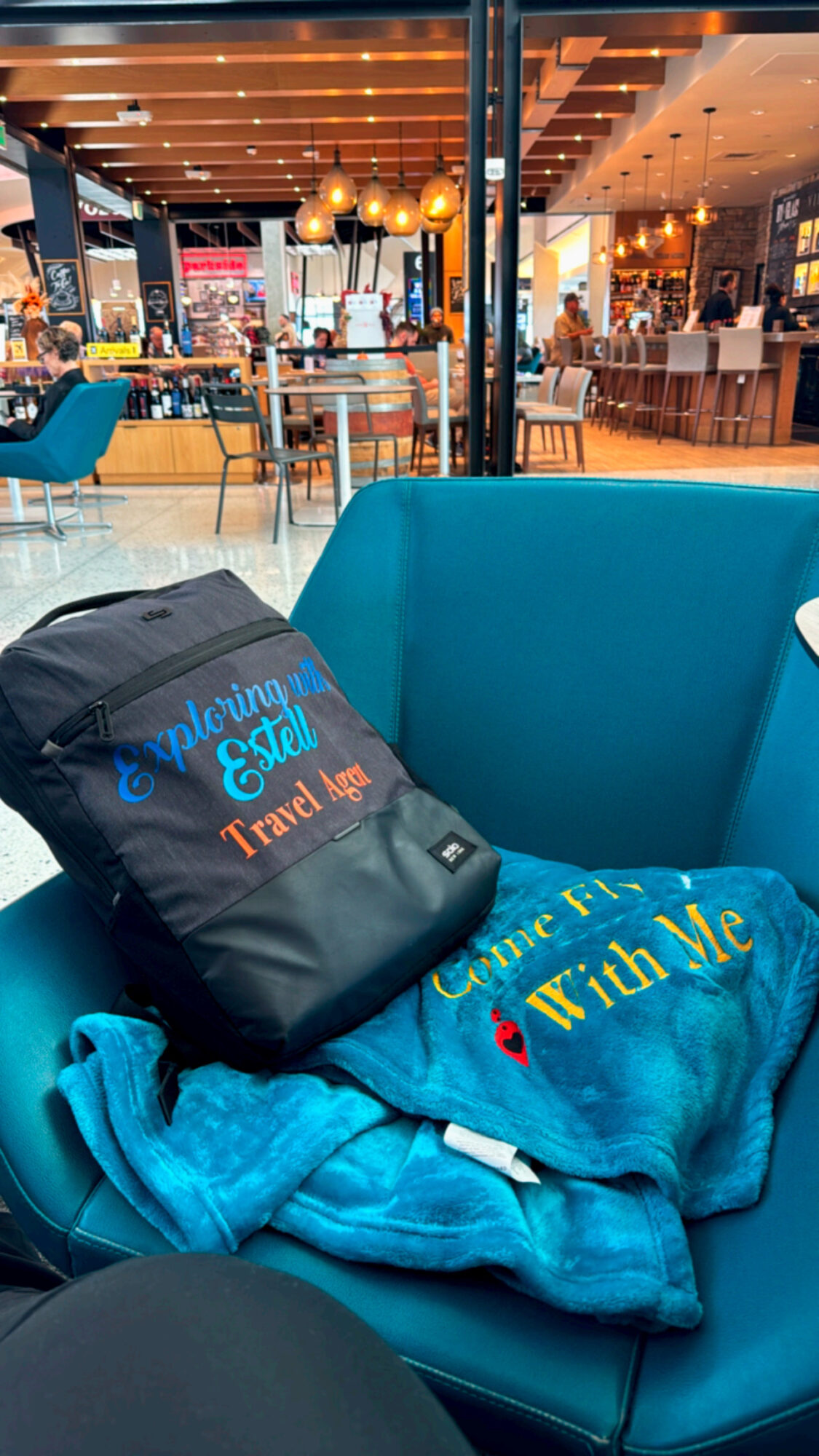 Indoor restaurant with seating, tables, and warm lighting, viewed through a window with a blue chair and backpack in foreground.
