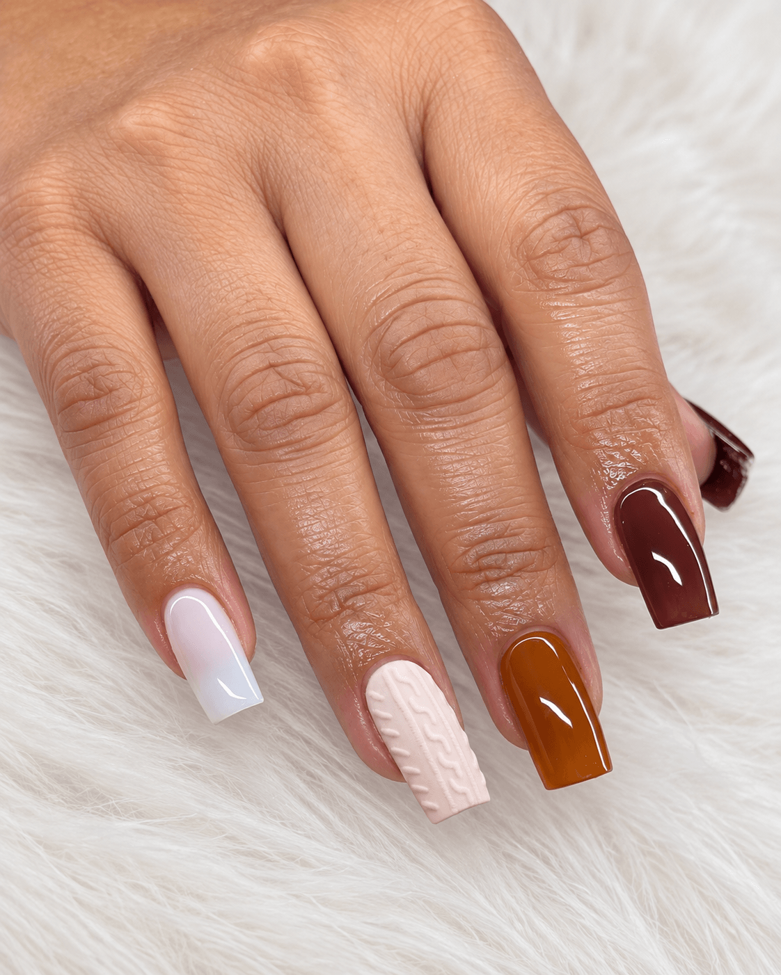 Hand with four nails painted in white, pink, brown, and dark red, resting on a soft surface.