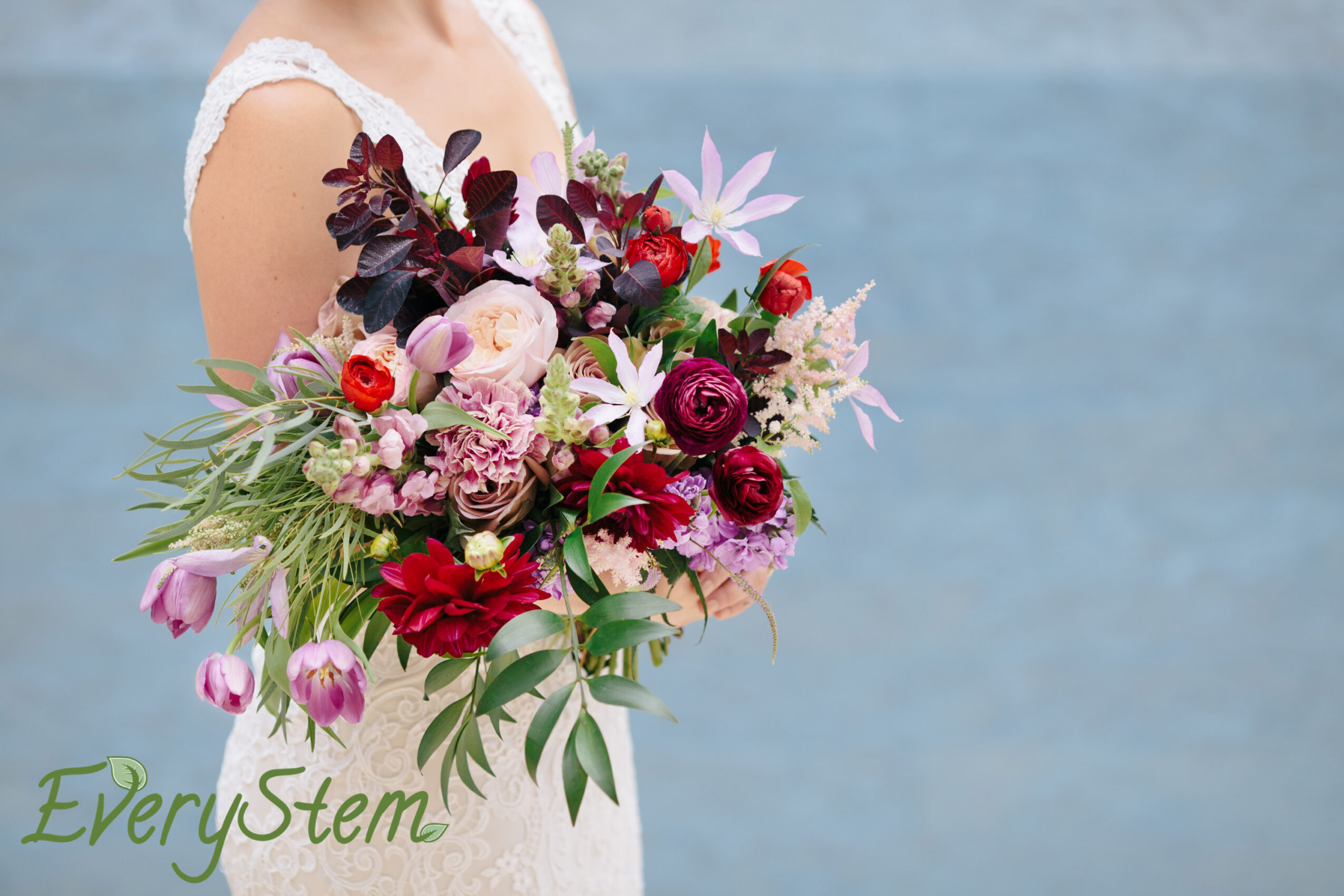Person holding a colorful bouquet of flowers against a blue background, wearing a white dress, with green text reading 'EveryStem'.