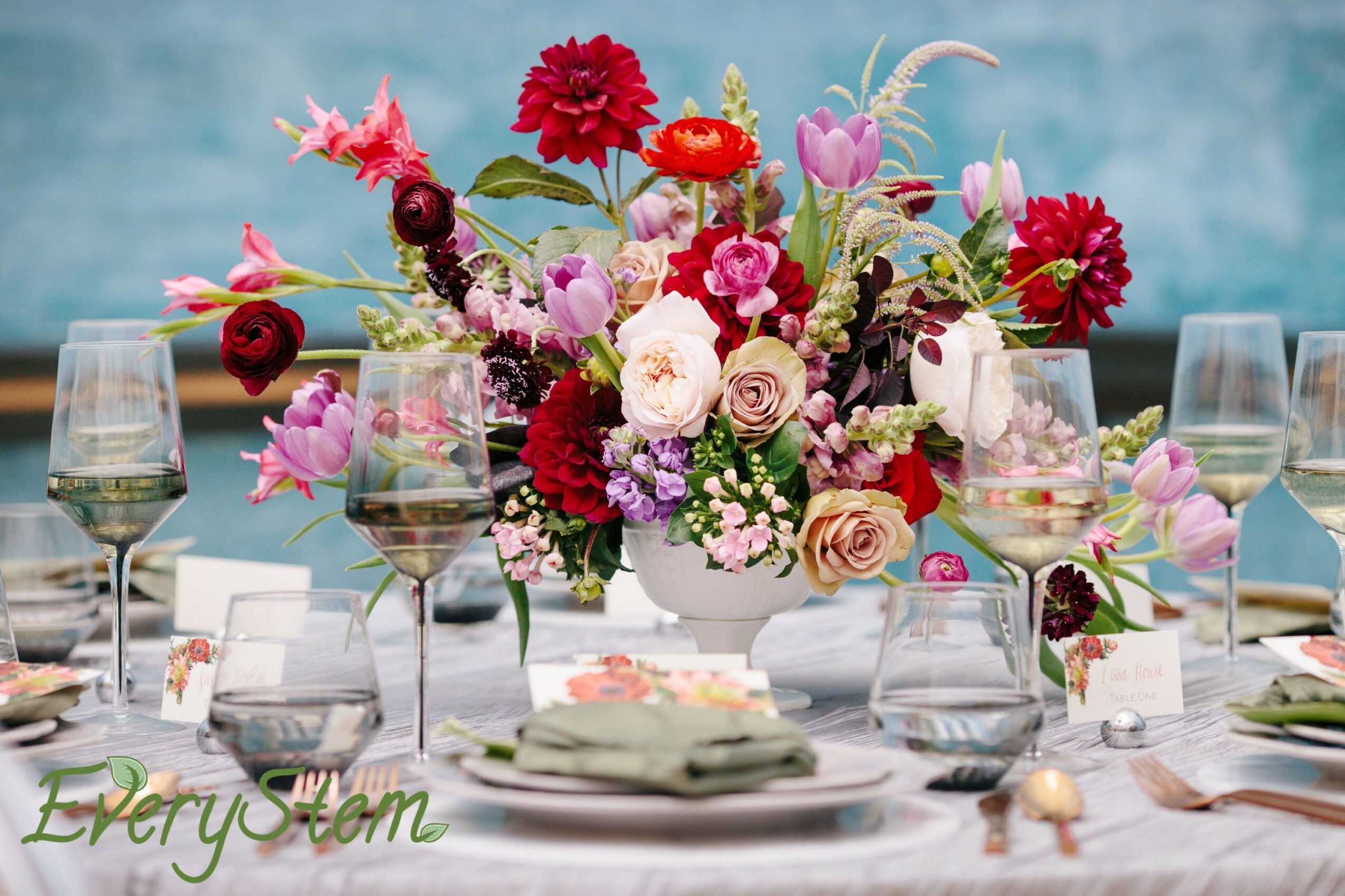 Table with a large colorful flower arrangement, glasses, plates, and napkins, set for a formal event, against a blue background.