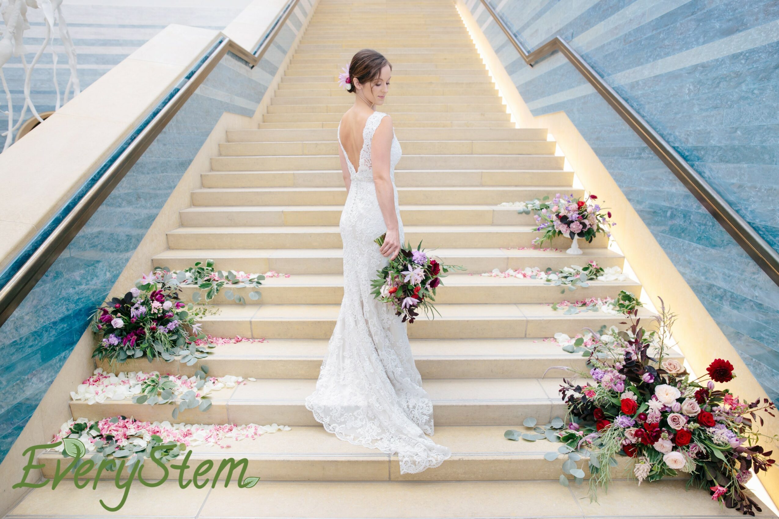 Woman in wedding dress holding bouquet on stairs with floral arrangements, glass building background.