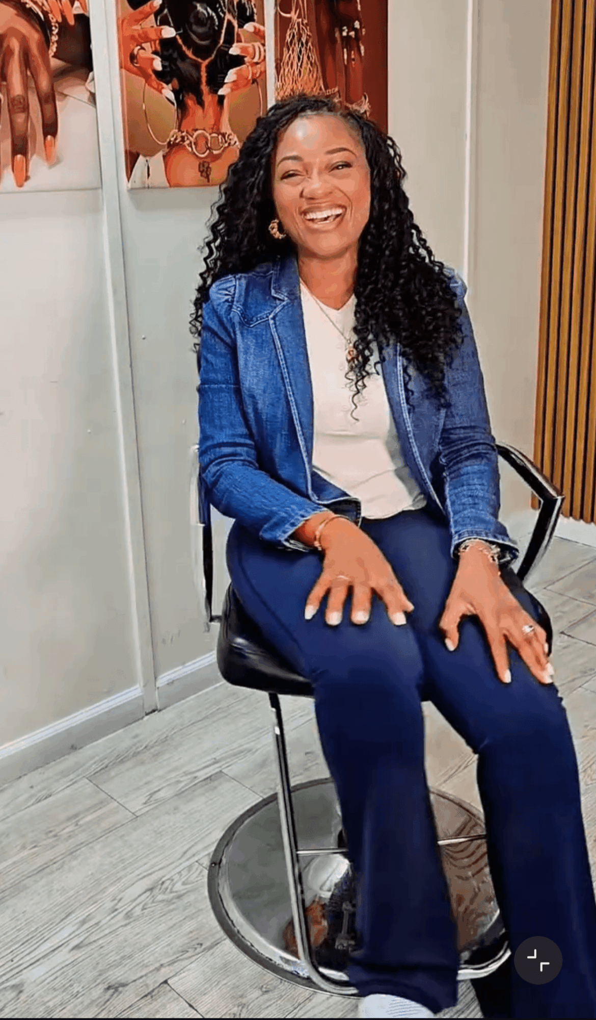 Smiling woman with curly hair sitting on a stool in a room with artwork and wooden paneling behind her.