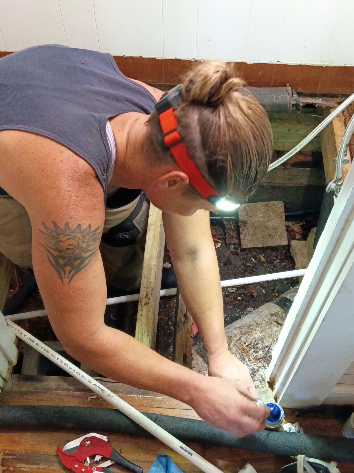 Person working on a construction or repair project, wearing a headlamp, with a tattoo on their arm, leaning over a window frame.