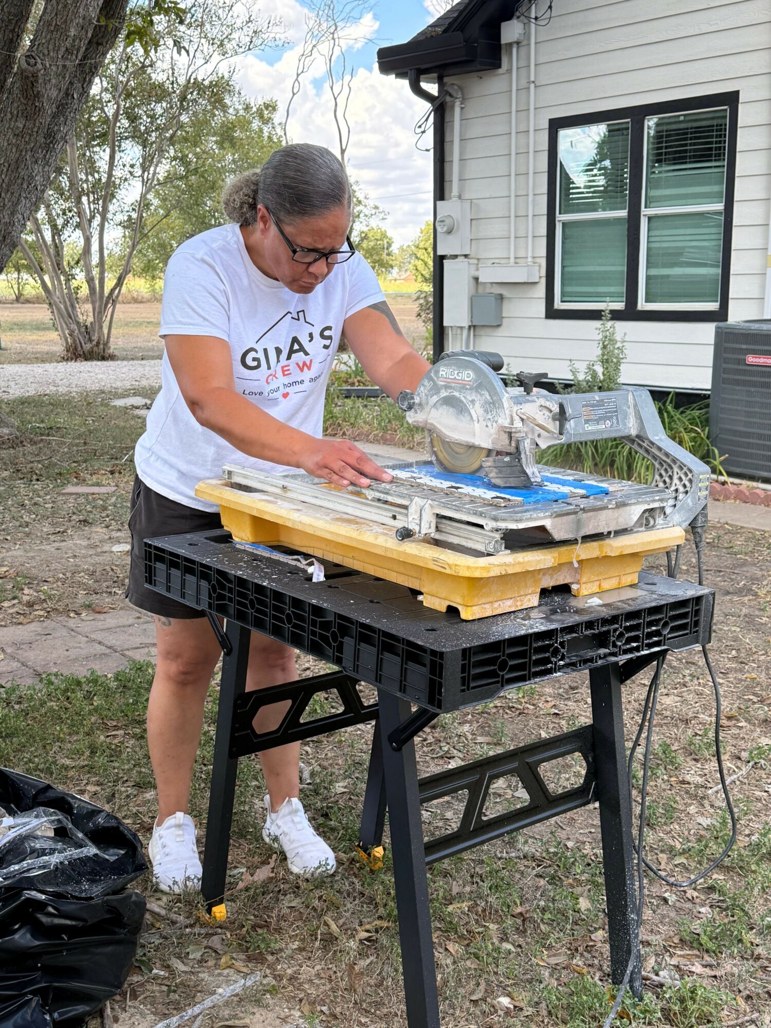 Person wearing glasses and a white T-shirt working on a project outdoors, with a house and trees in the background.