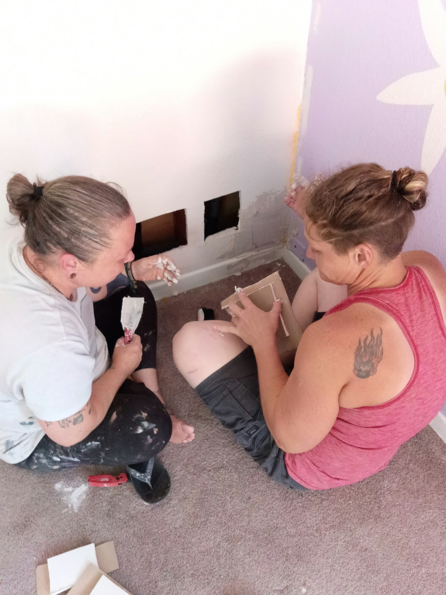 Two women sitting on the floor, one applying paint to the wall, the other holding a paintbrush and a container.