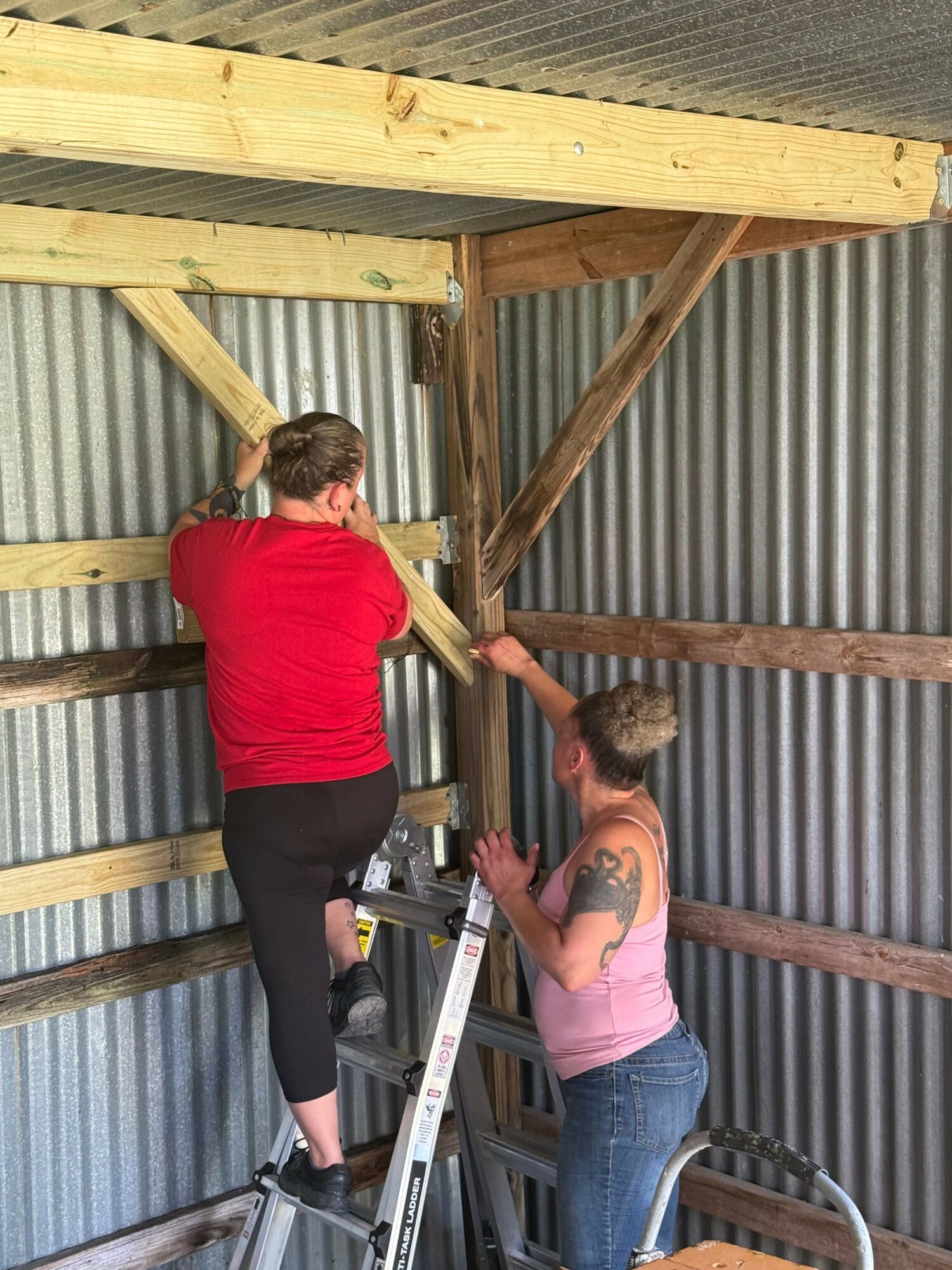 Two women working on a construction project, one in a pink top and the other in a red top, with wooden beams and metal roofing visible.