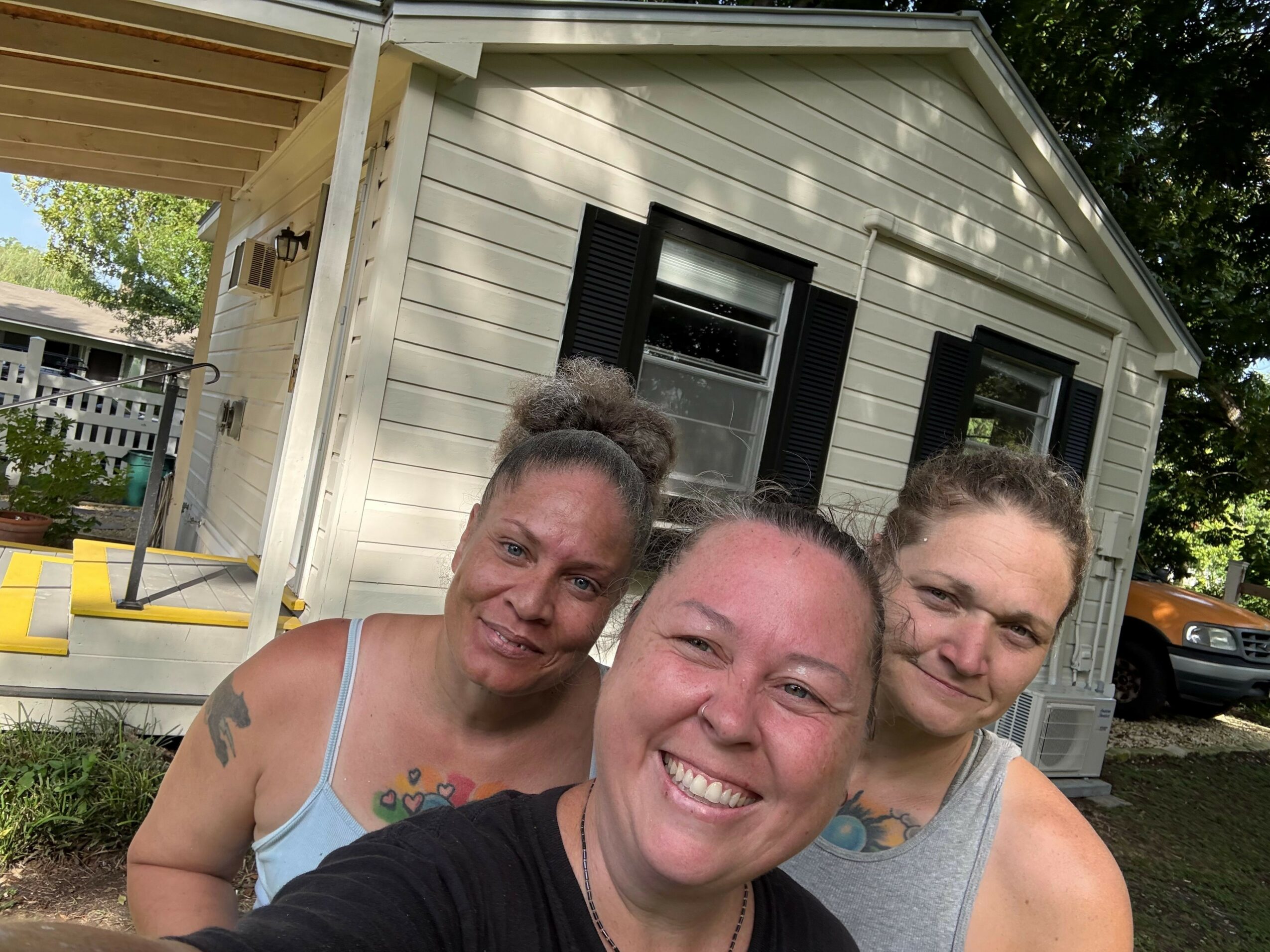 Three smiling people taking a selfie outside a house with white siding and black shutters.