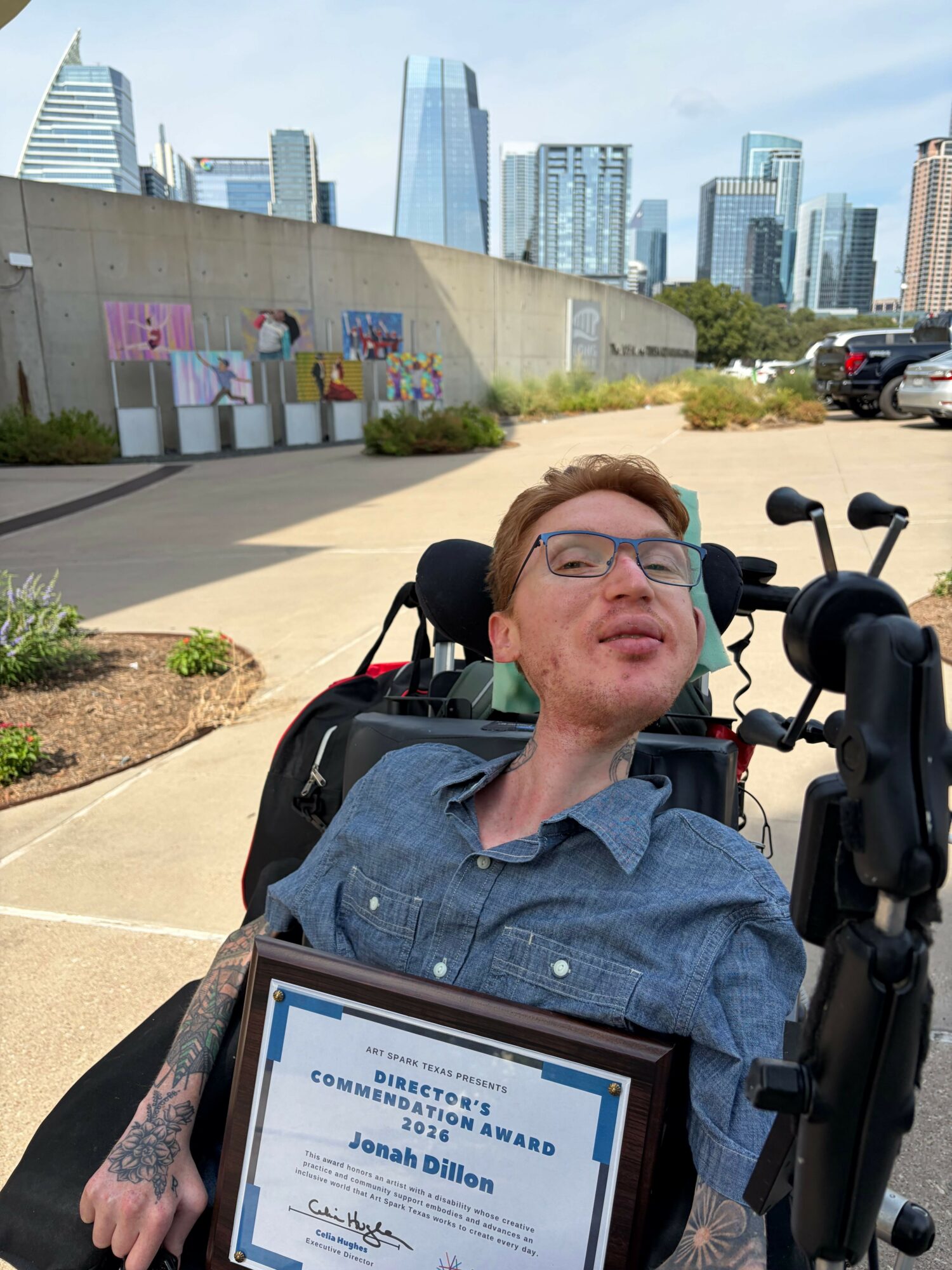 Person in wheelchair holding a framed certificate outdoors with city buildings in background.