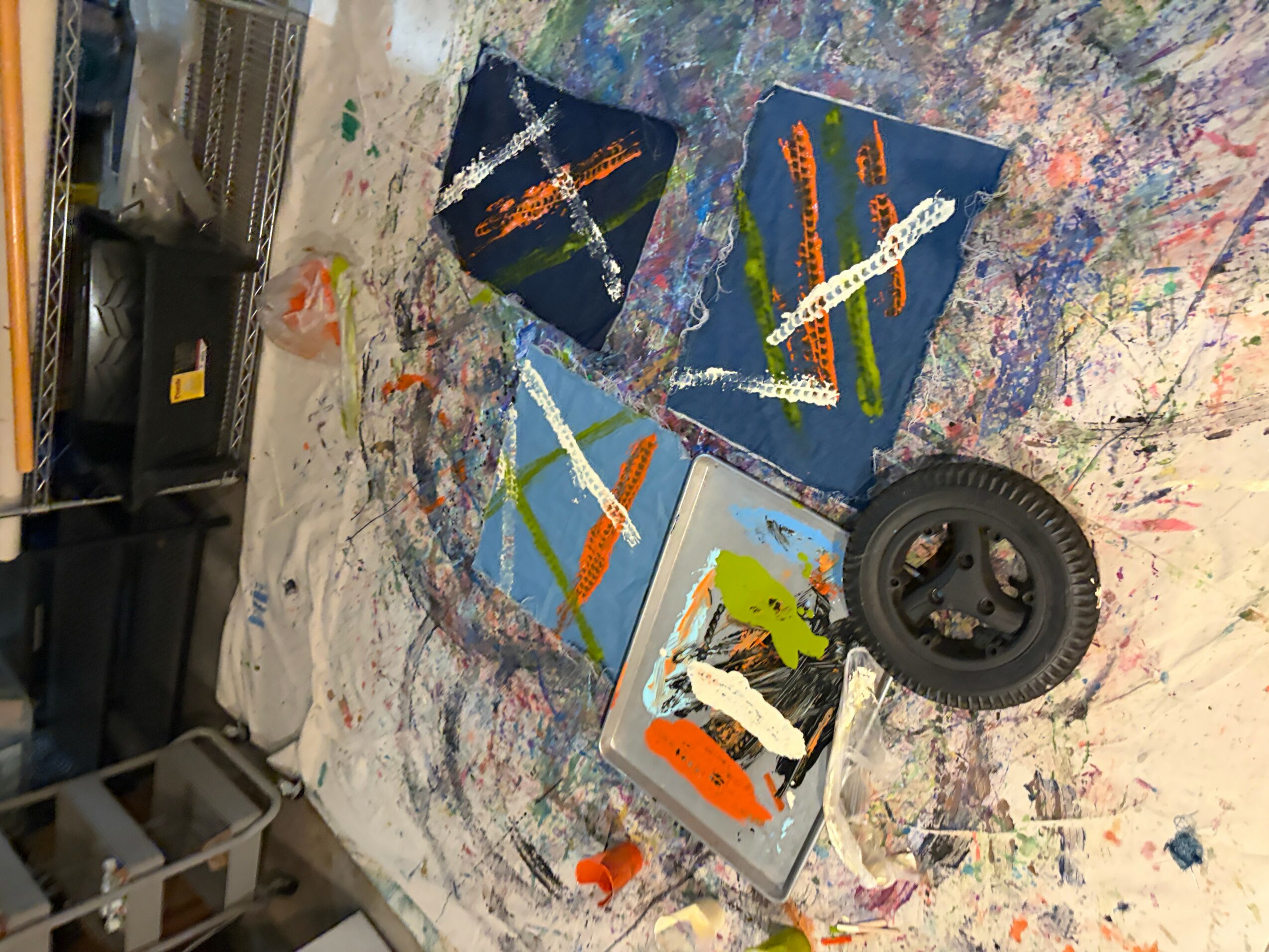 Paintings on a table with paint splatters, a black wheel, and art supplies, viewed from the side.