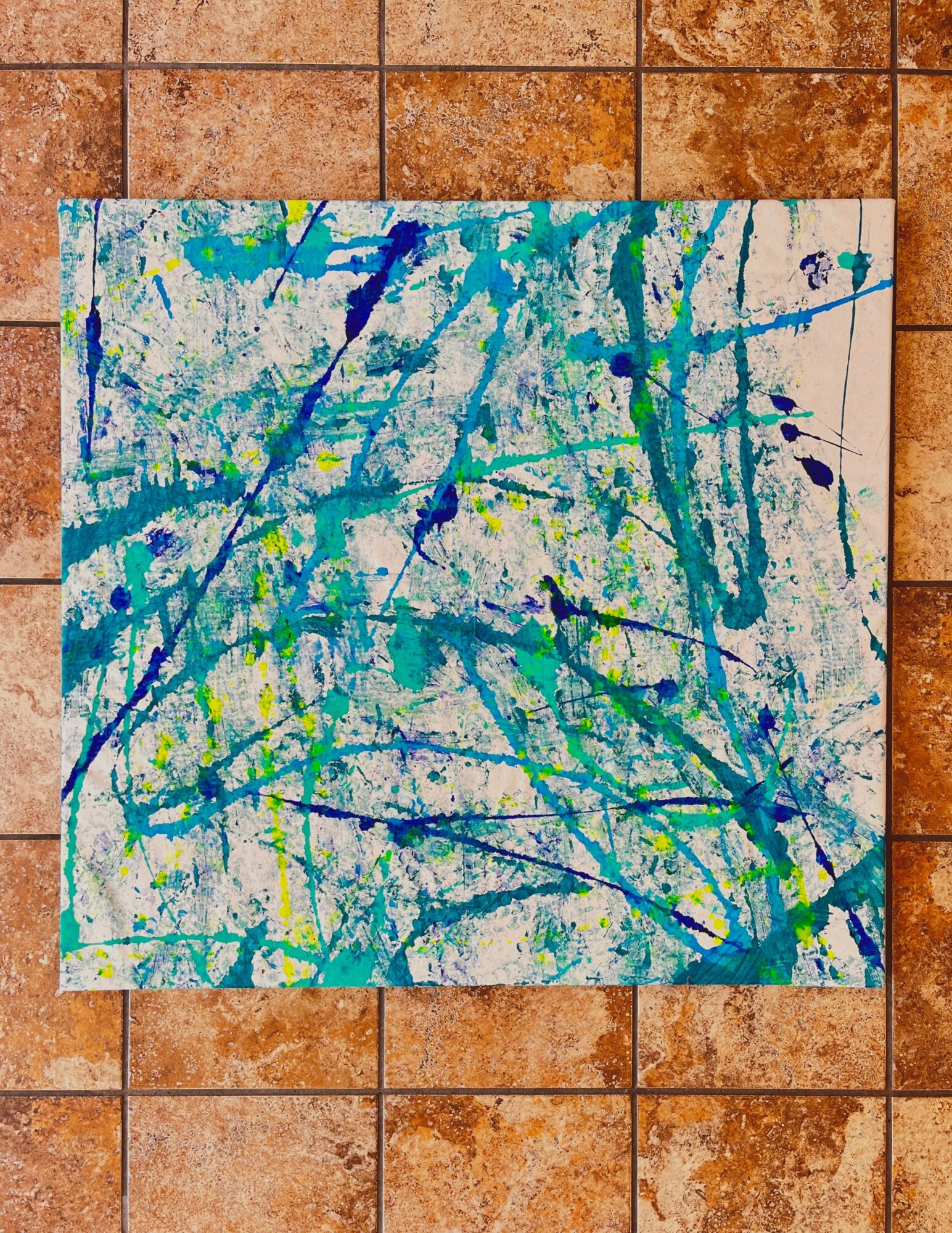 Abstract painting with blue, green, and yellow splatters on a white background, placed on a tiled floor.