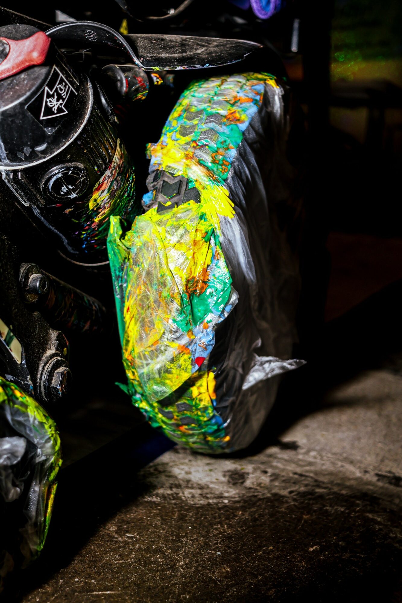 Motorcycle tire with colorful paint splatters, partially covered by a black helmet, on a dark surface.