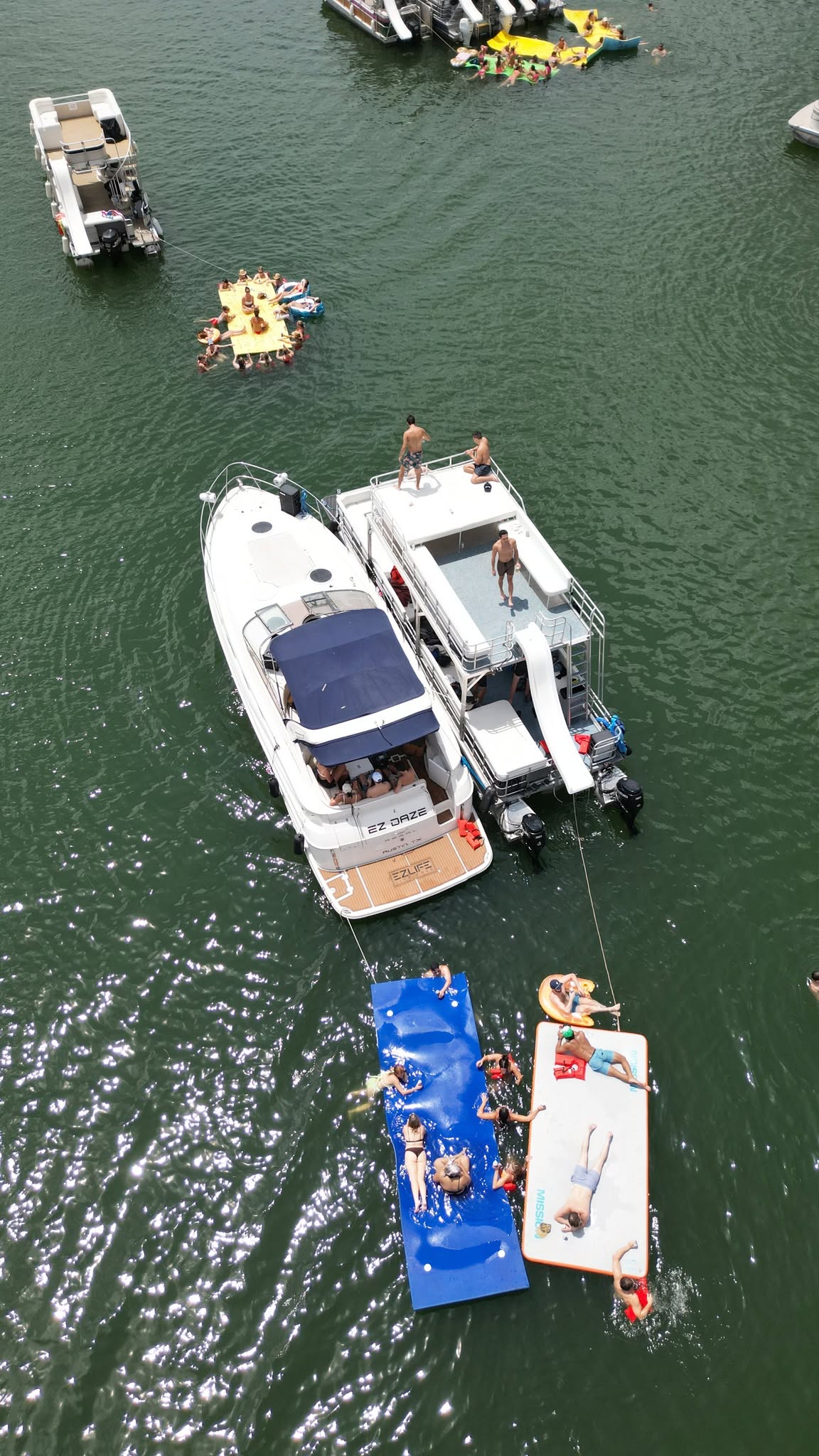 Multiple boats and floating platforms on water, with people relaxing and enjoying the day.