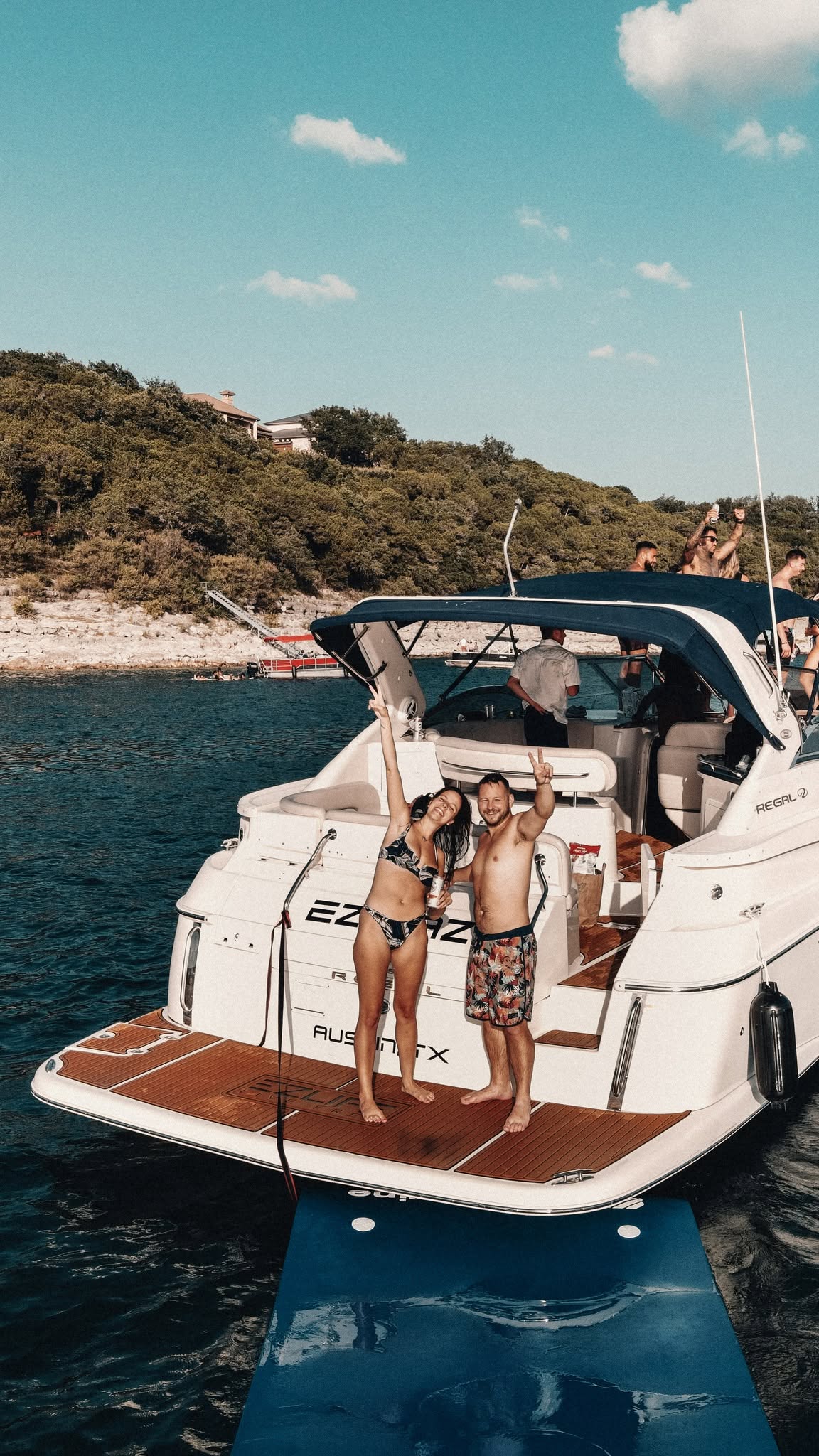 Two children in swimsuits stand on the back of a white boat, holding a rope, with water and a hillside in the background.