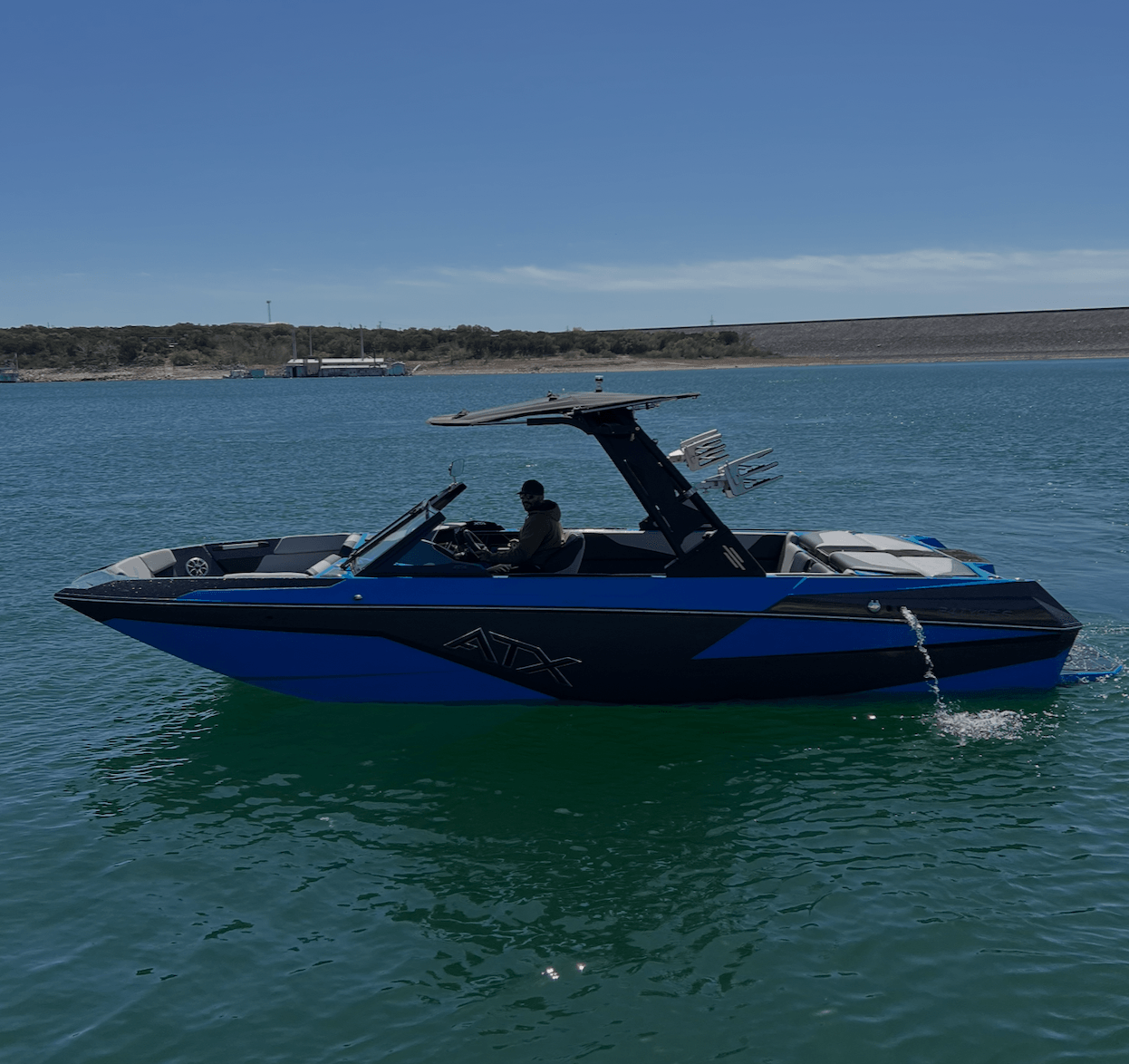 Motorboat on water with person inside, canopy open, shoreline in background, clear sky.