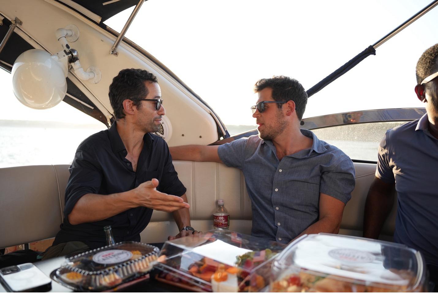 Three men sitting on a boat, engaged in conversation, with food and drinks on the table in front of them.