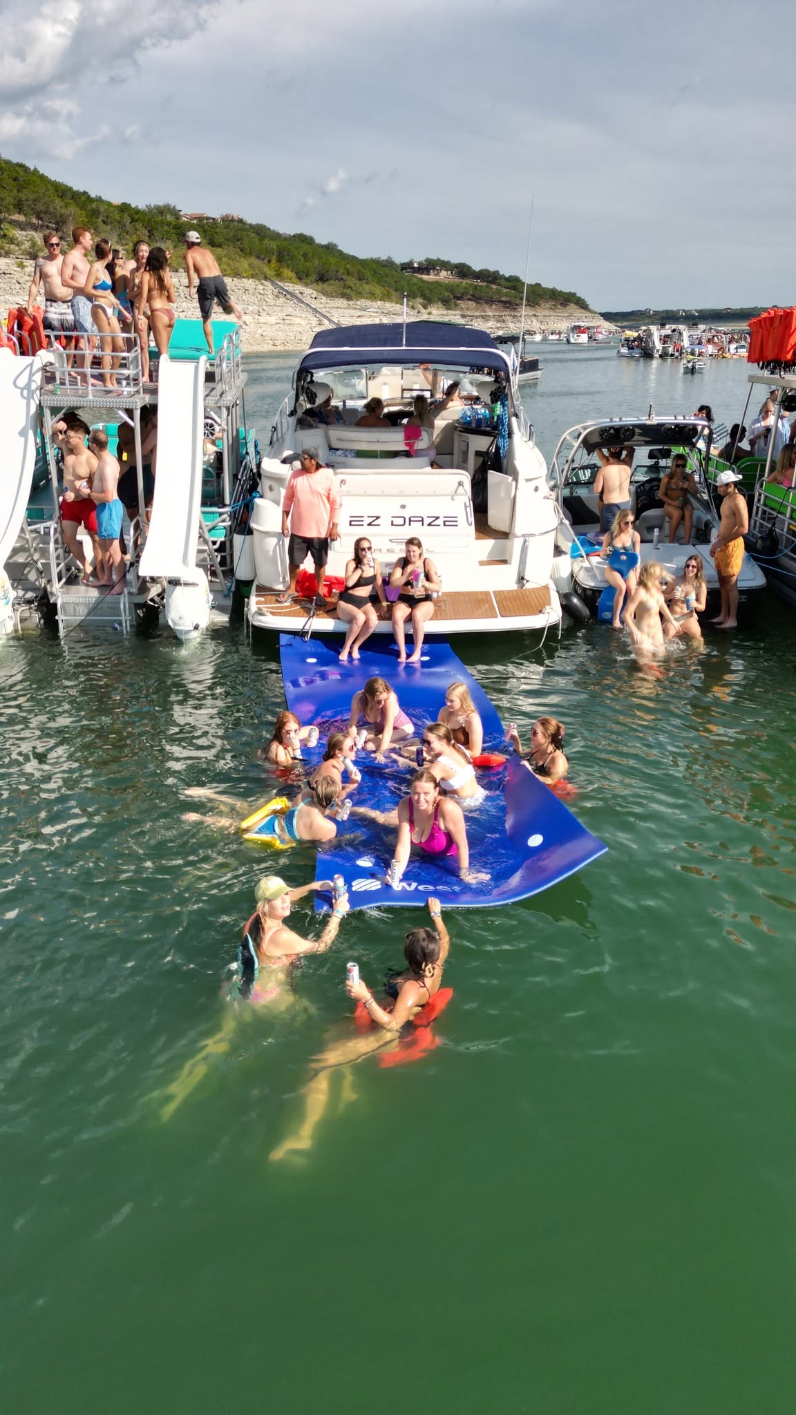 People swimming and relaxing near a boat docked at a marina with boats and a shoreline in the background.